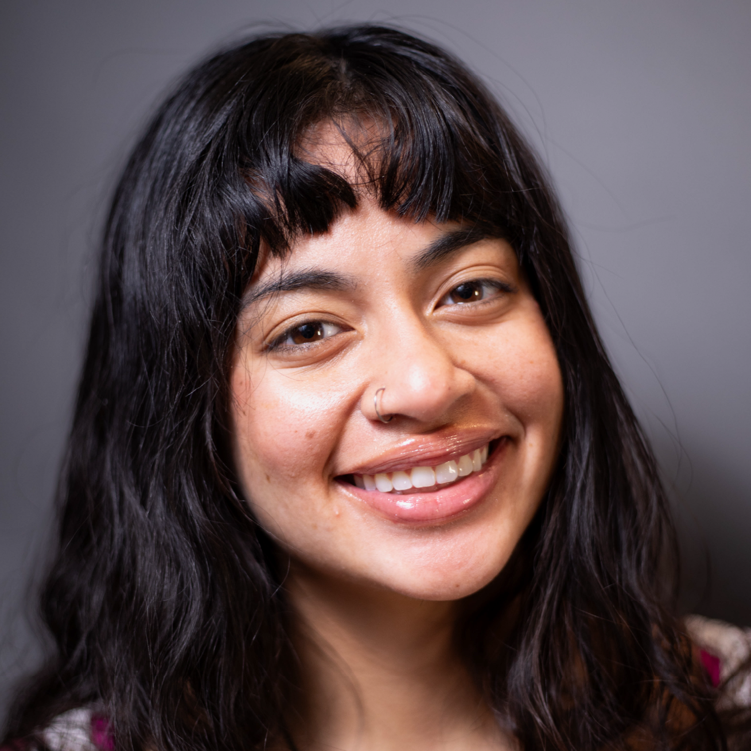 Close-up of a young woman with black wavy hair and bangs, smiling against a gray background.