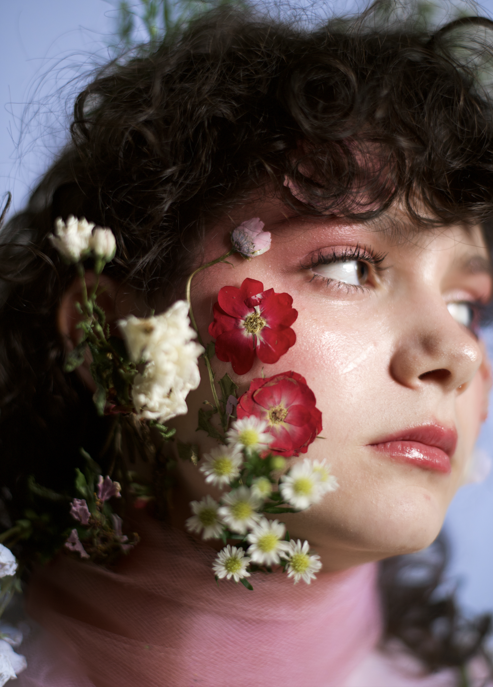 Close-up of a person with curly hair, flowers on his face, and a blue background.