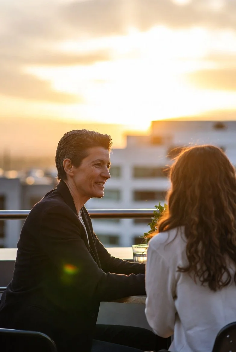 A Middle Eastern mentor and South Asian mentee smile warmly at a rooftop café with a wrapped gift on the table between them
