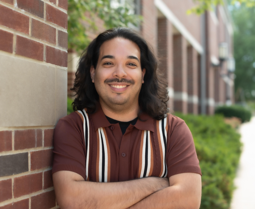 A smiling man with shoulder-length dark hair, a mustache, wearing a navy blue collared shirt, standing indoors with sunlight and greenery visible in the background.