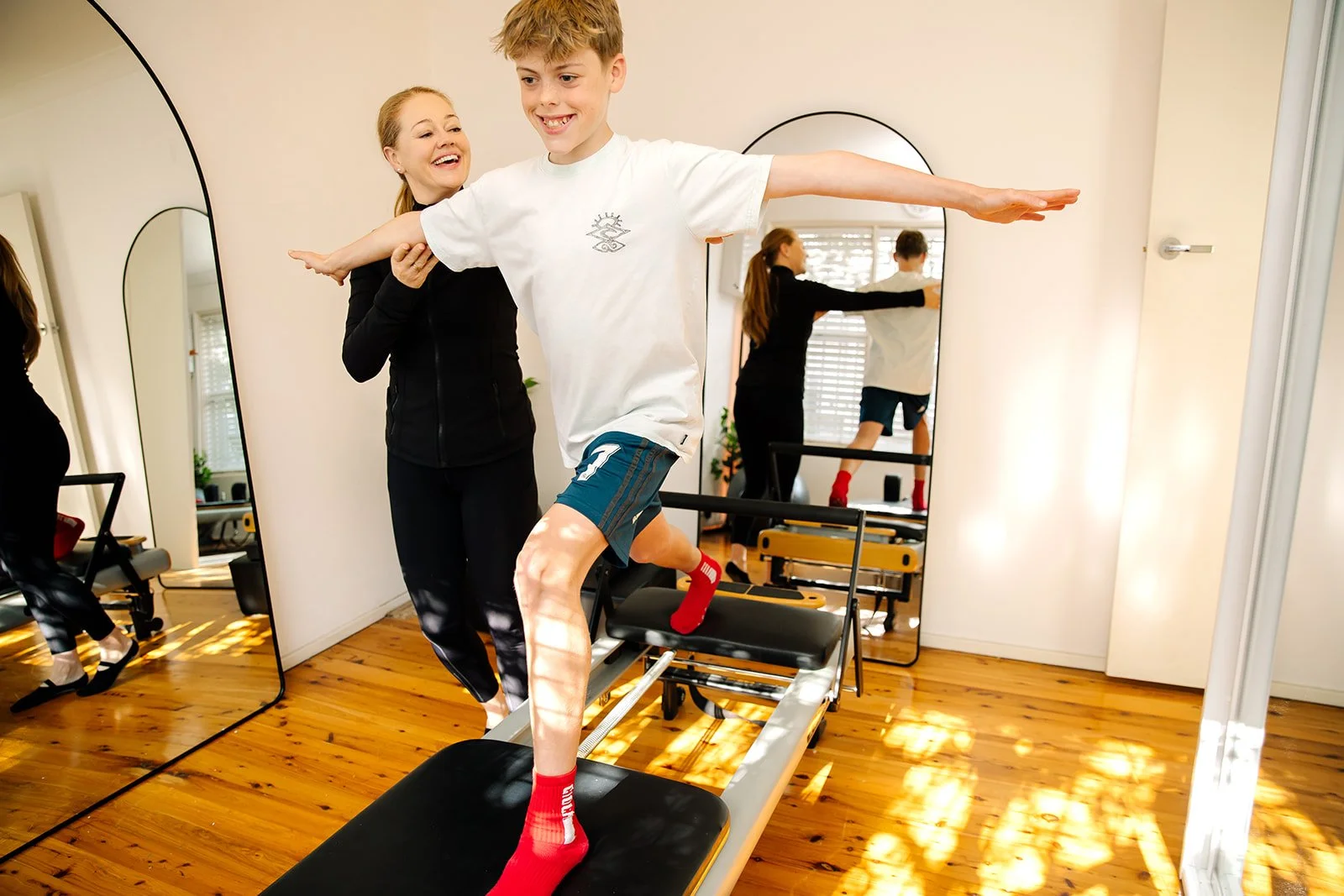 A young boy in workout clothes balancing on a treadmill with a woman behind him assisting, in a room with wooden floors and a large mirror.