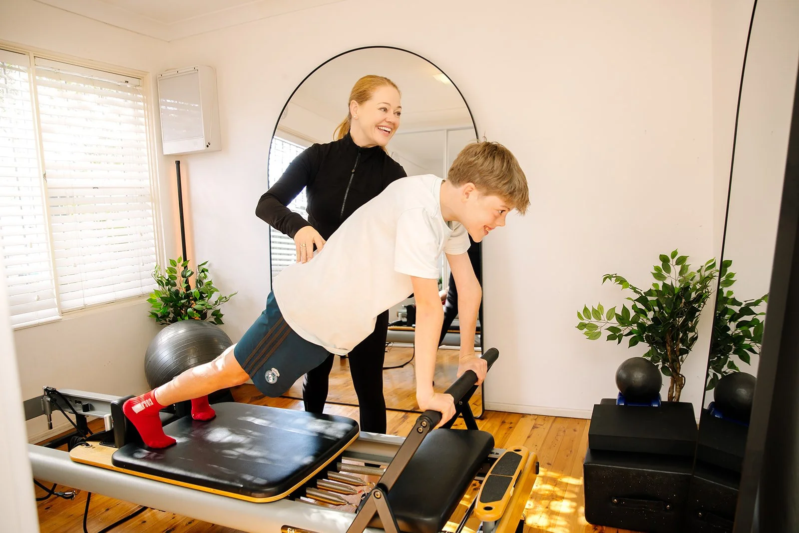Clinical Pilates performing a plank exercise on a Pilates reformer in a bright room with wooden floors, plants, and a round mirror.