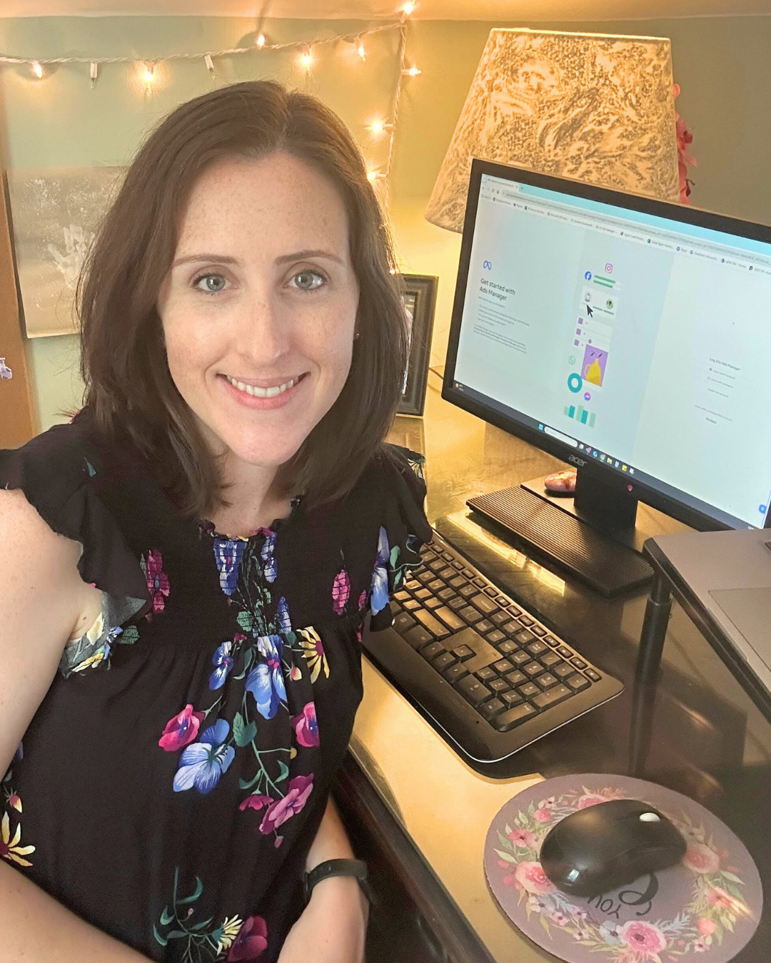 A woman with shoulder-length brown hair, smiling, sitting at a desk with a computer monitor, keyboard, and mouse.