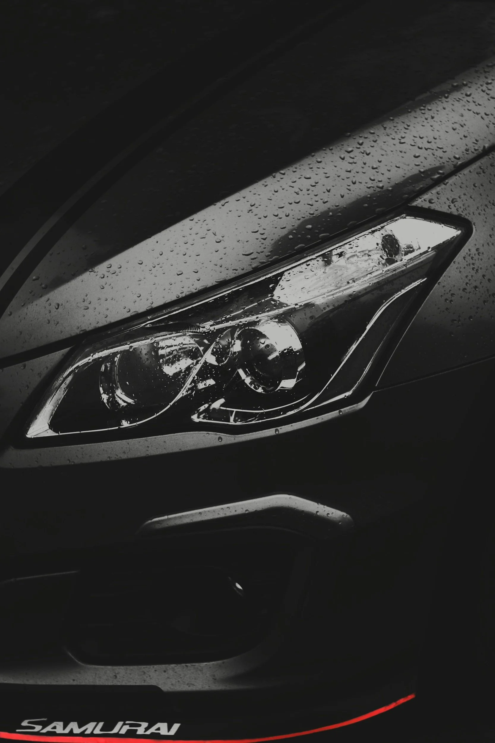 Close-up of a black car headlight with raindrops on the surface, and the word 'SAMURAI' written in white and red at the bottom.