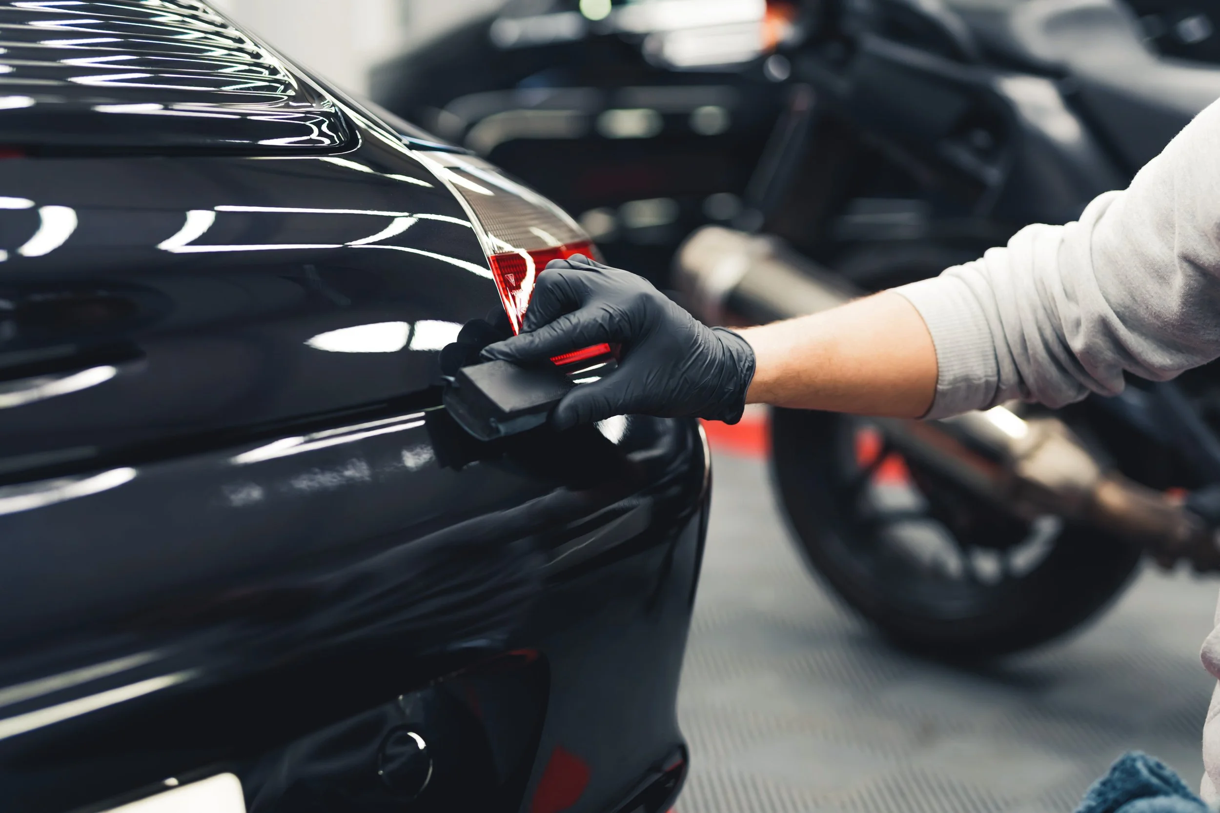 A person wearing a black glove using a detailing tool to polish the rear of a black car, with a motorcycle in the background.