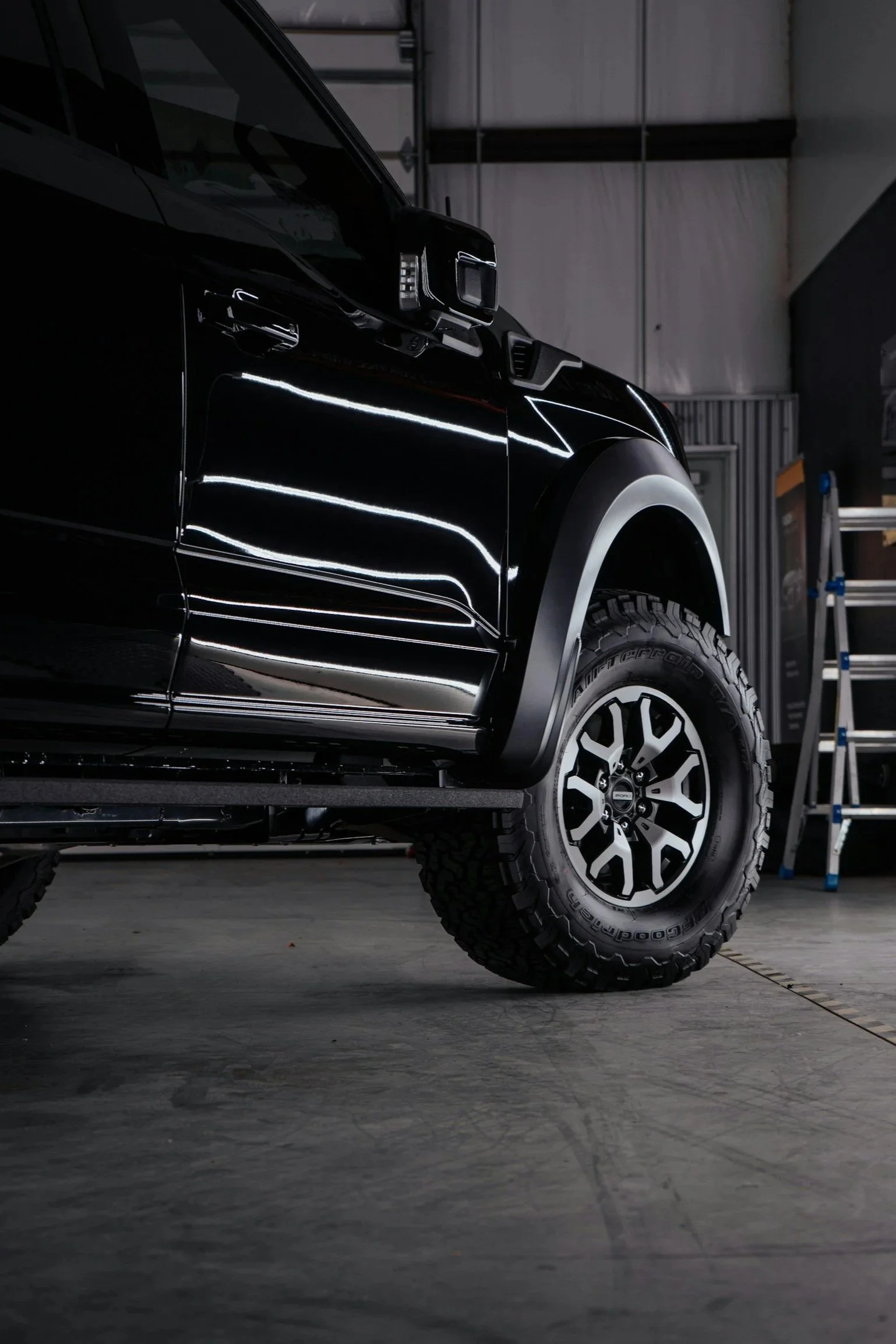 Close-up of a black off-road vehicle's front side, showing rugged tire and mirror, inside a garage with a ladder in the background.