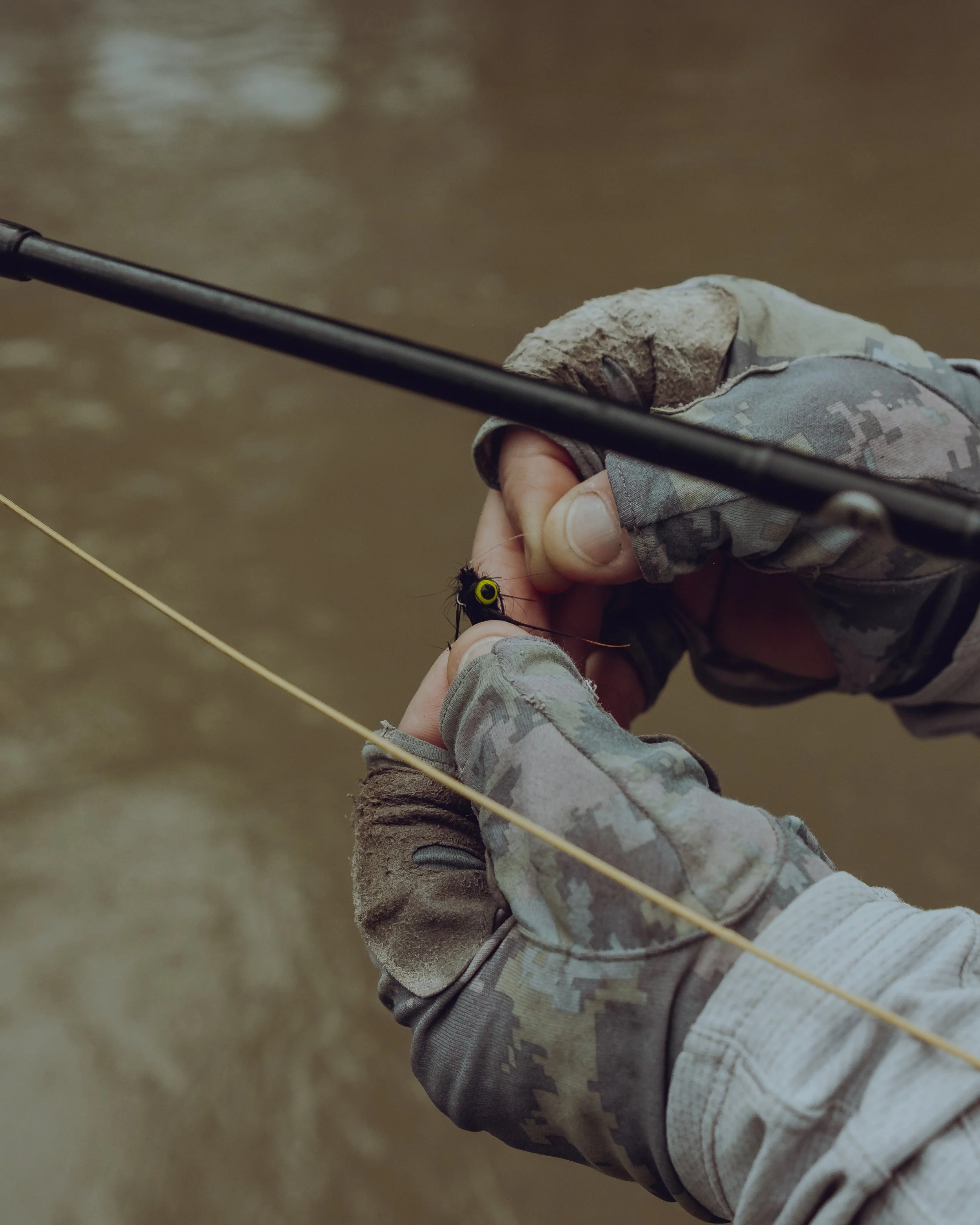 Person wearing camouflage gloves tying a fishing hook on a line by a river.