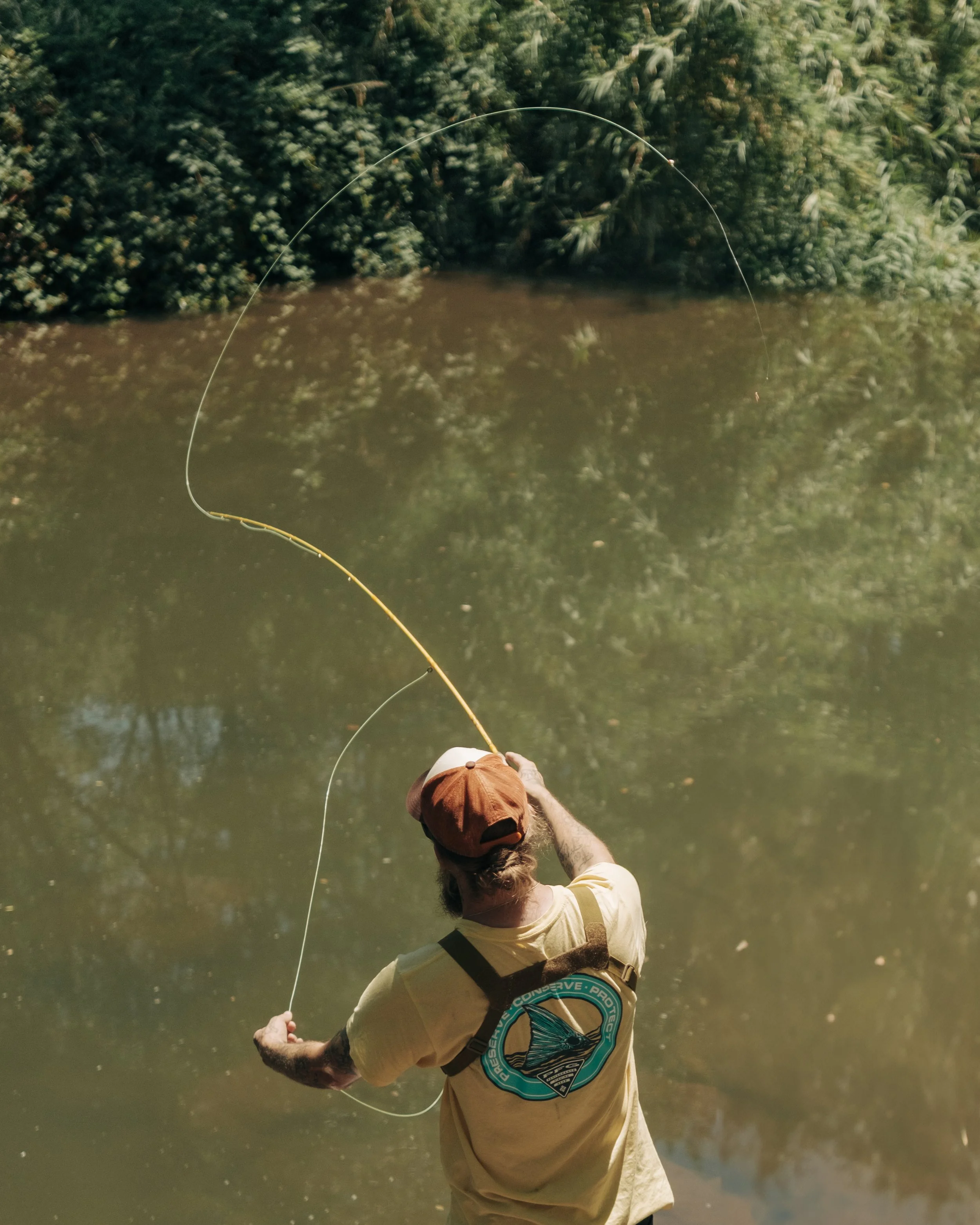 A man stands by a river, casting a fishing line into the water surrounded by lush green foliage.