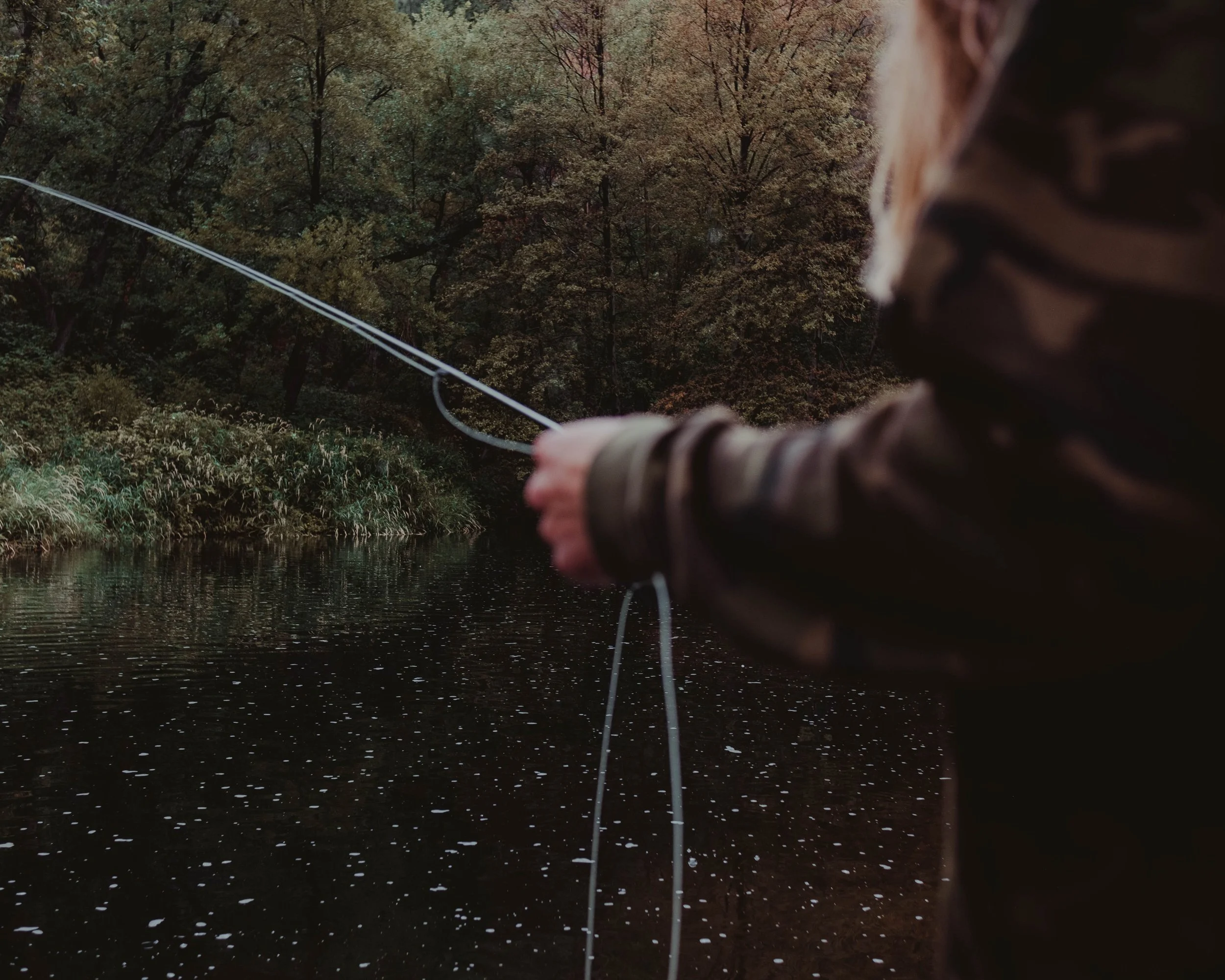 A person fishing in a river or lake surrounded by trees with fall foliage, seen from behind, wearing a camouflage jacket.
