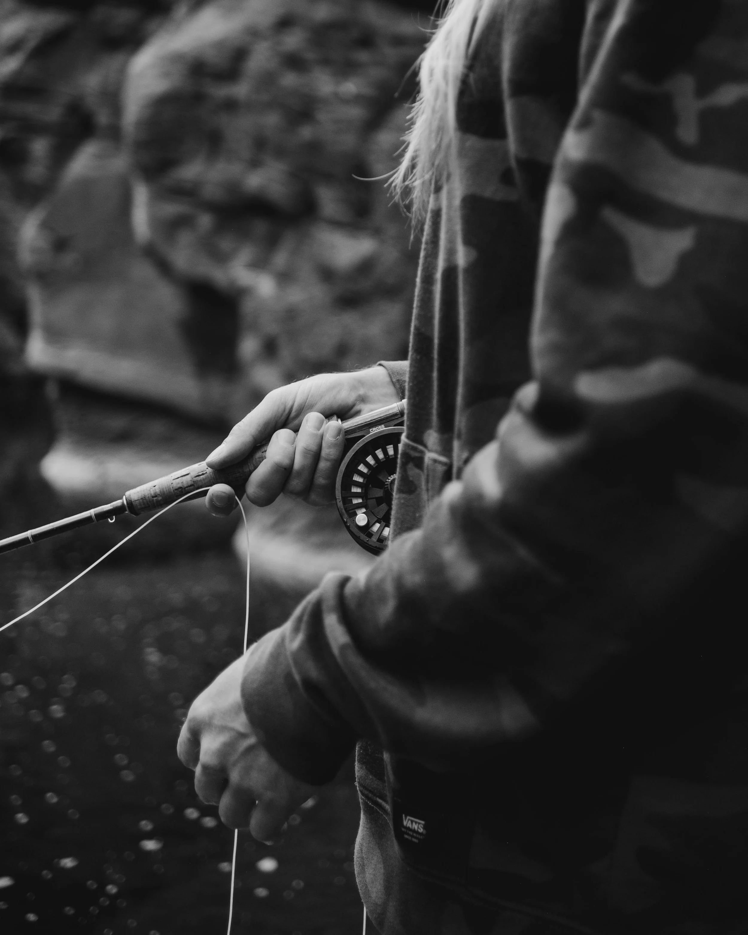 A person fishing with a fly rod near a body of water, wearing a camouflage jacket.