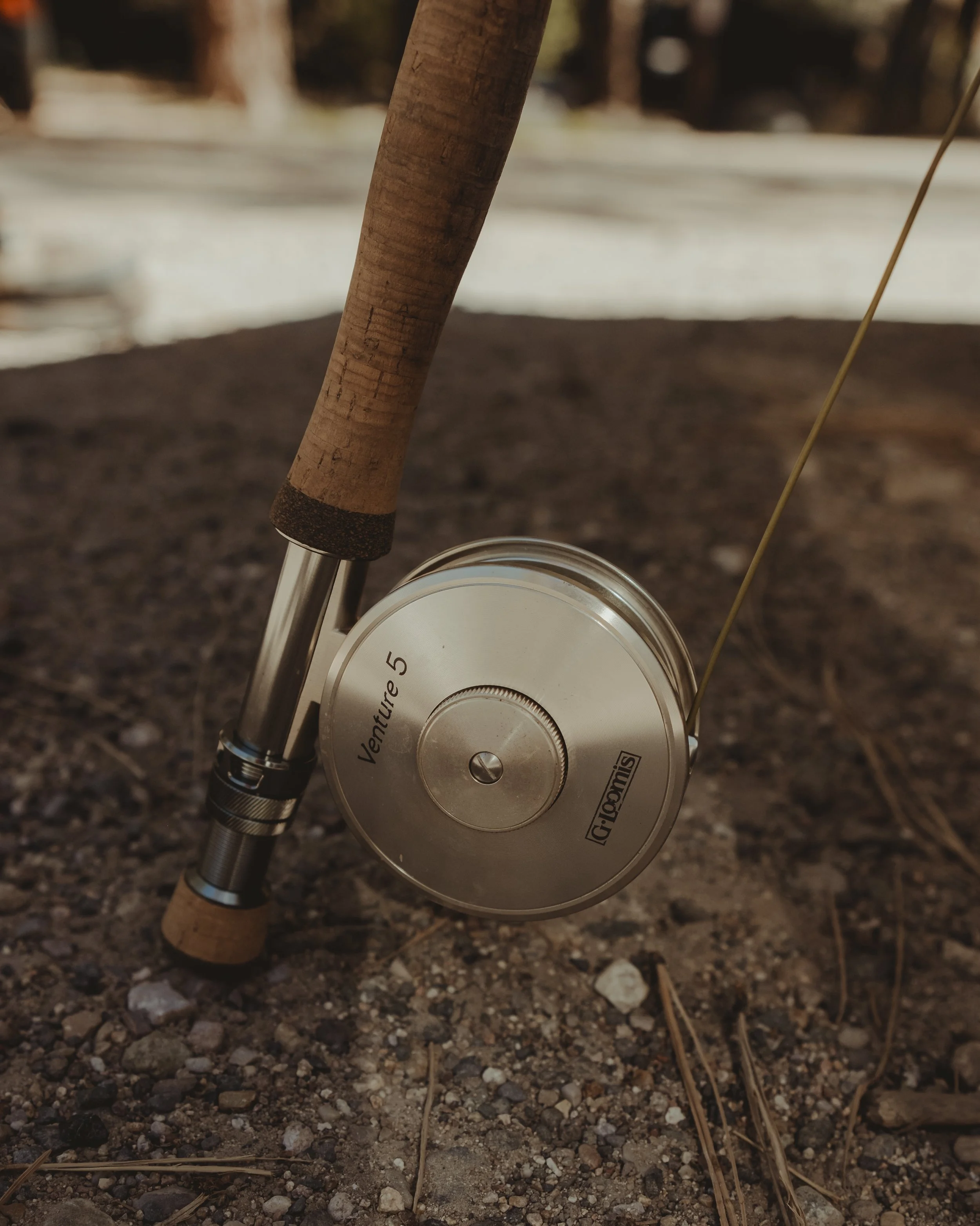 Close-up of a fishing reel labeled 'Venture 5' attached to a fishing rod, resting on gravelly ground outdoors.