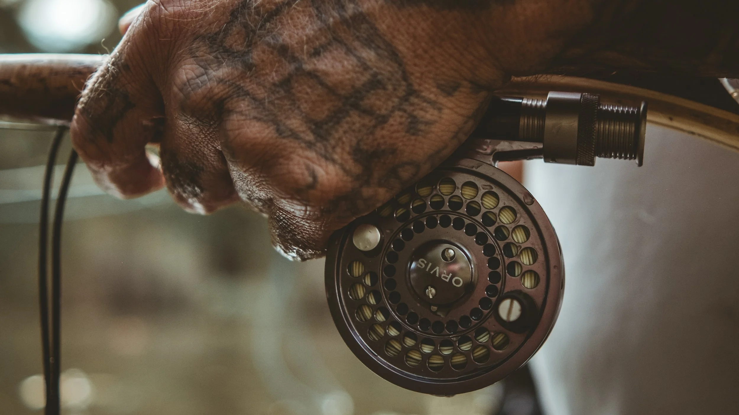 A hand with tattoos holding a vintage fly fishing reel with a black handle, resting on a wooden surface.