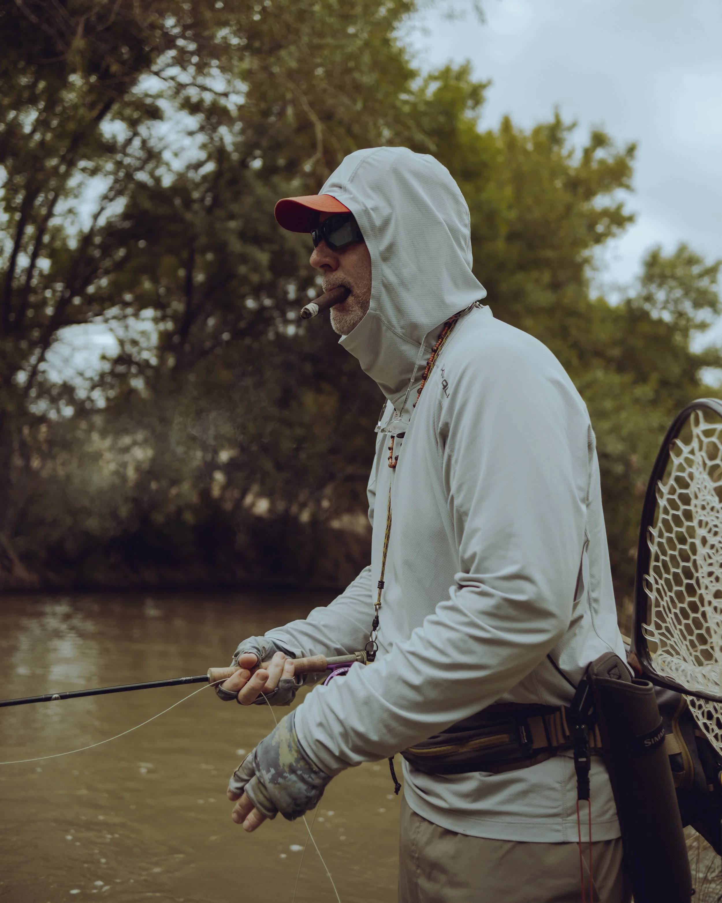 Man fishing in a river, wearing a white hoodie, sunglasses, and a hat, smoking a cigar, with trees in the background.