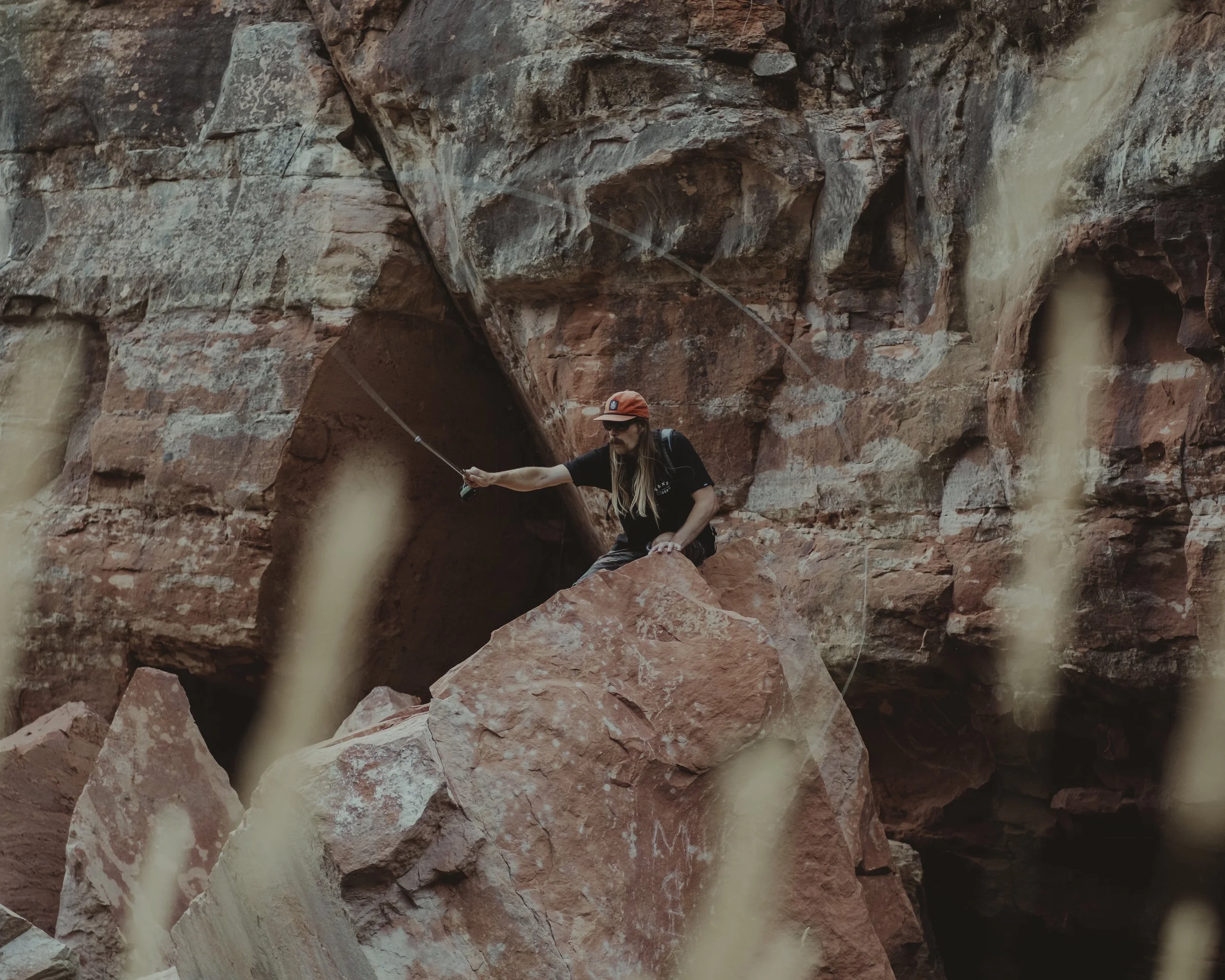 A woman with long hair, sunglasses, and a red cap sitting on a large rock in a canyon, fishing with a fishing rod.