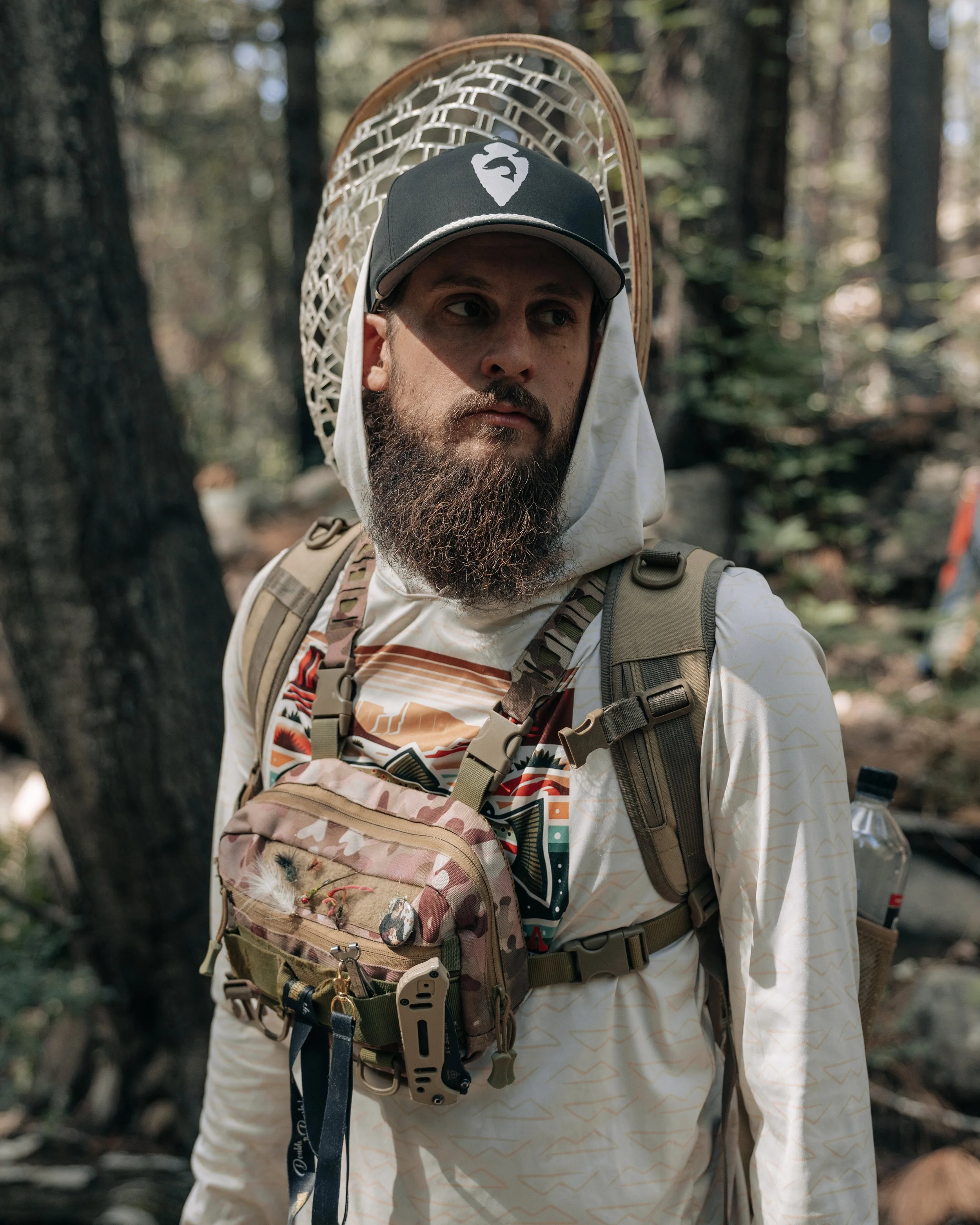 A man with a beard wearing a camouflage backpack, a cap, and a net hat, standing in a forest.