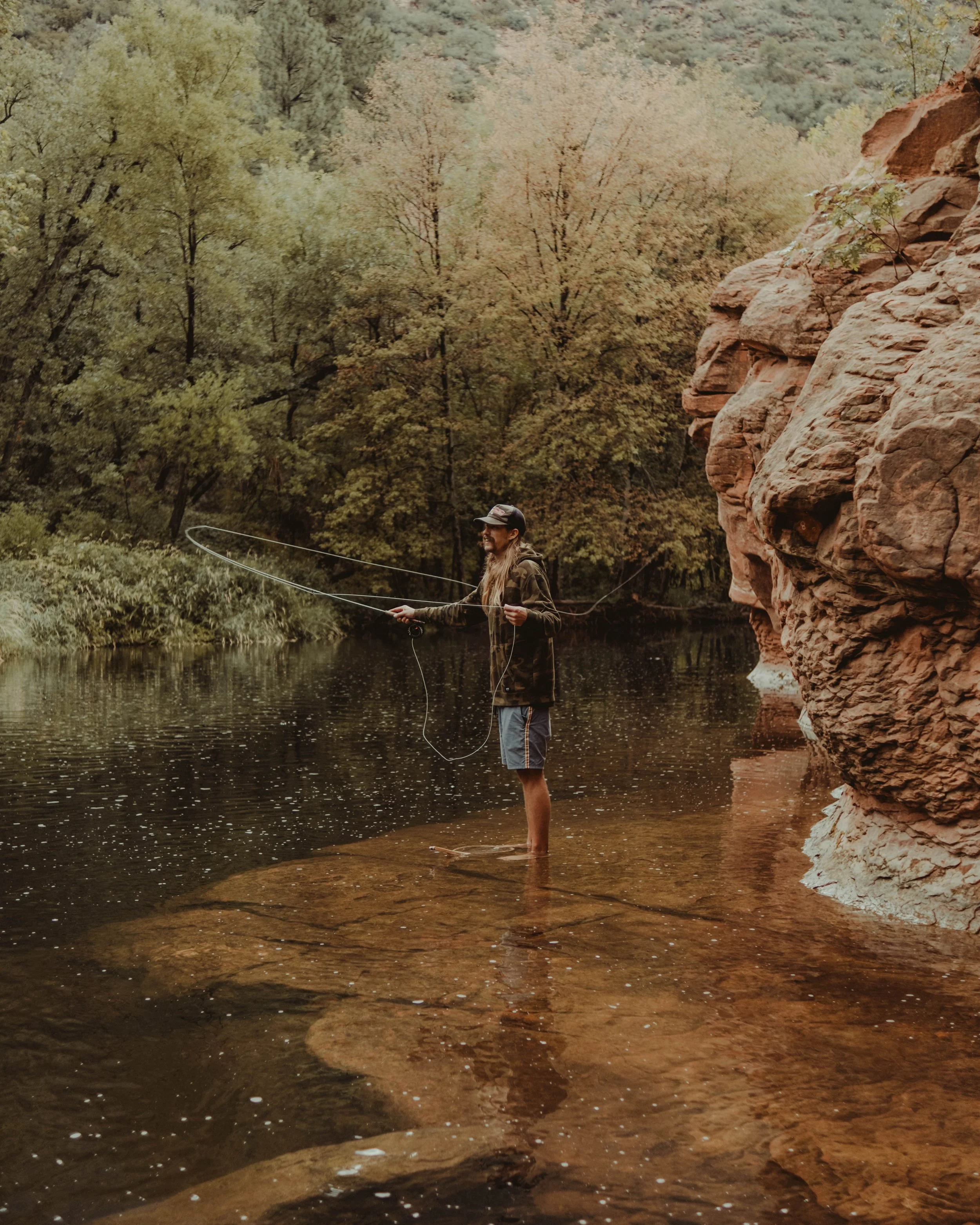 Person fishing in a river surrounded by trees and rocky cliffs with autumn-colored leaves.