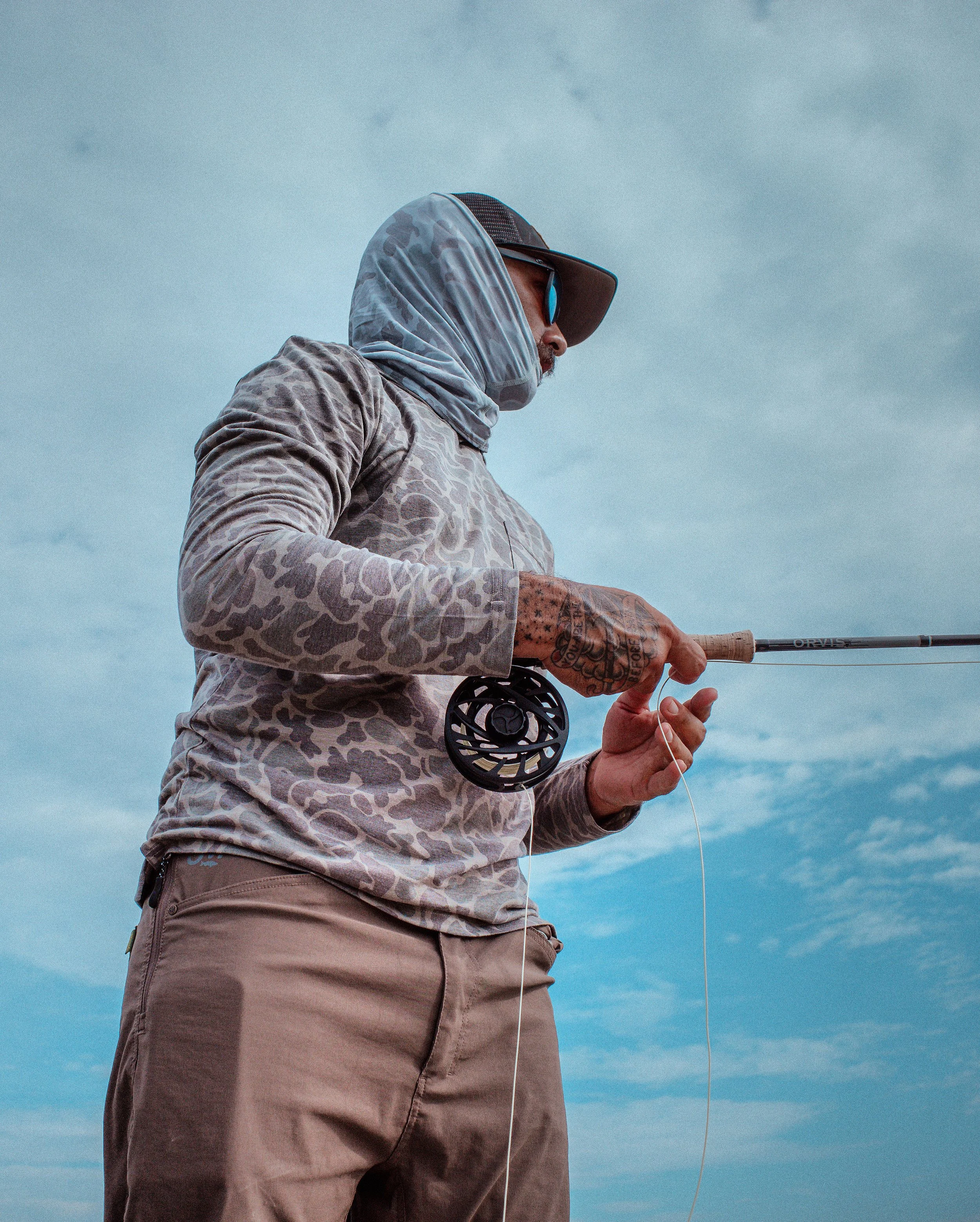 A man fishing outdoors, wearing sunglasses, a cap with a neck cover, and a long-sleeved shirt, holding a fishing rod against a cloudy sky.