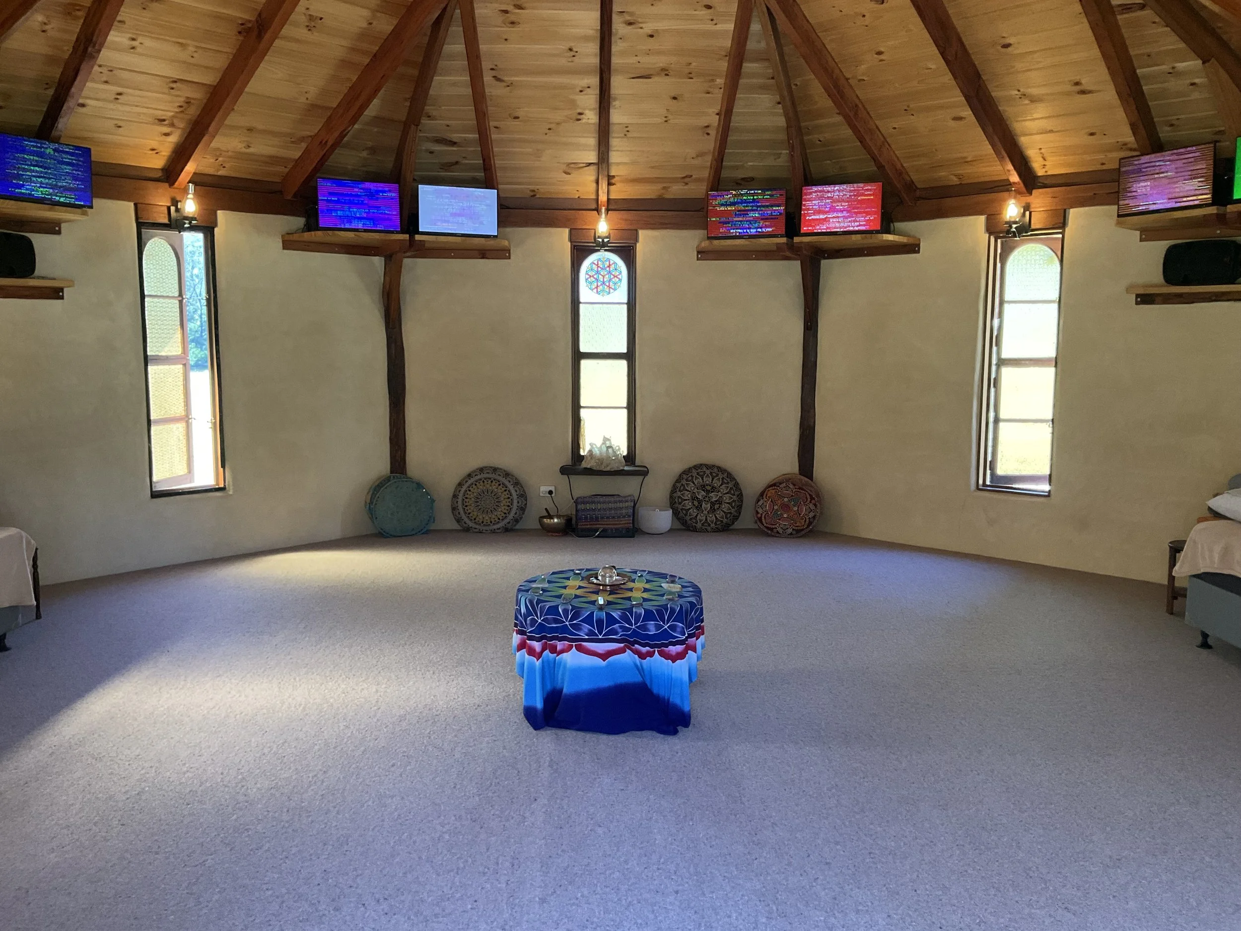 A spacious room with a wooden vaulted ceiling, three narrow stained glass windows, and five EESystem units mounted on the upper walls. There is a small table with a cloth in the center, with sunlight streaming through the windows.