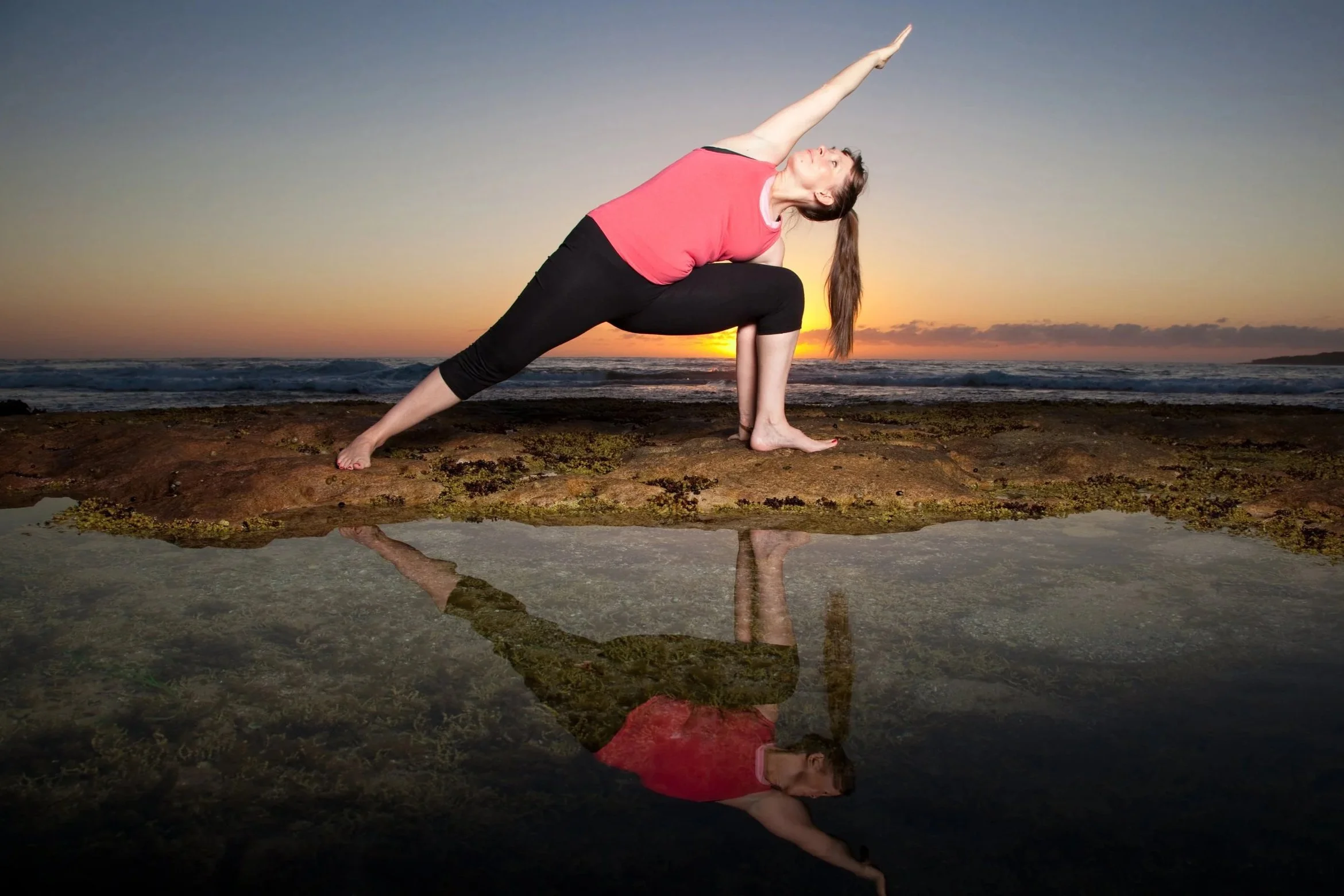 Tara practicing yoga on a rocky beach during sunrise, performing a side angle pose with her reflection visible in a small water pool.