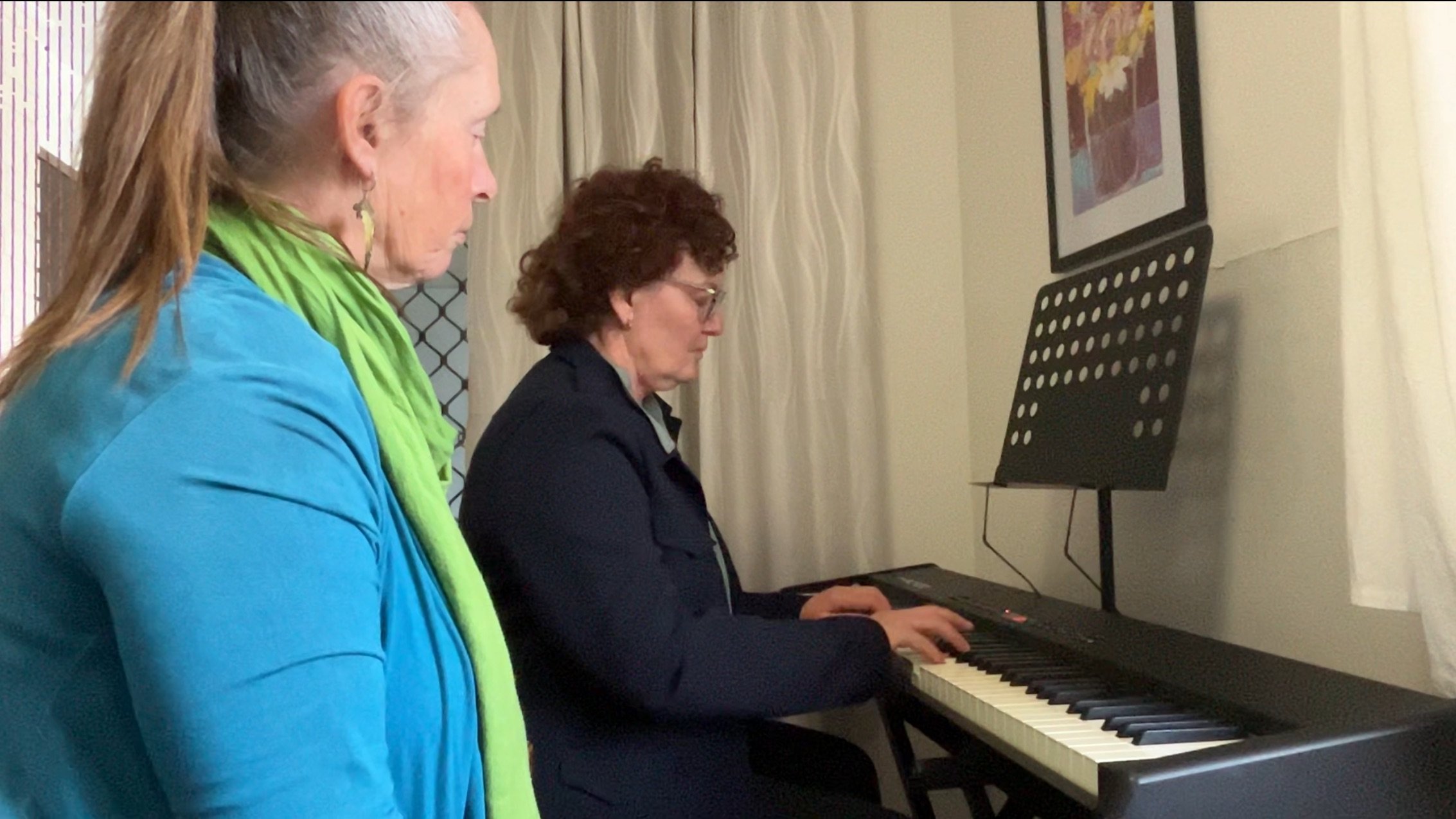 Two women are watching a woman playing a digital piano in a room with beige walls and artwork on the wall.