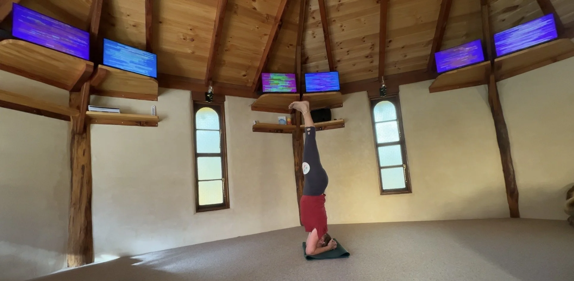 Tara practicing a headstand yoga pose in a room, the Hemple, with wooden beams and 6 EESystem units mounted on the ceiling.