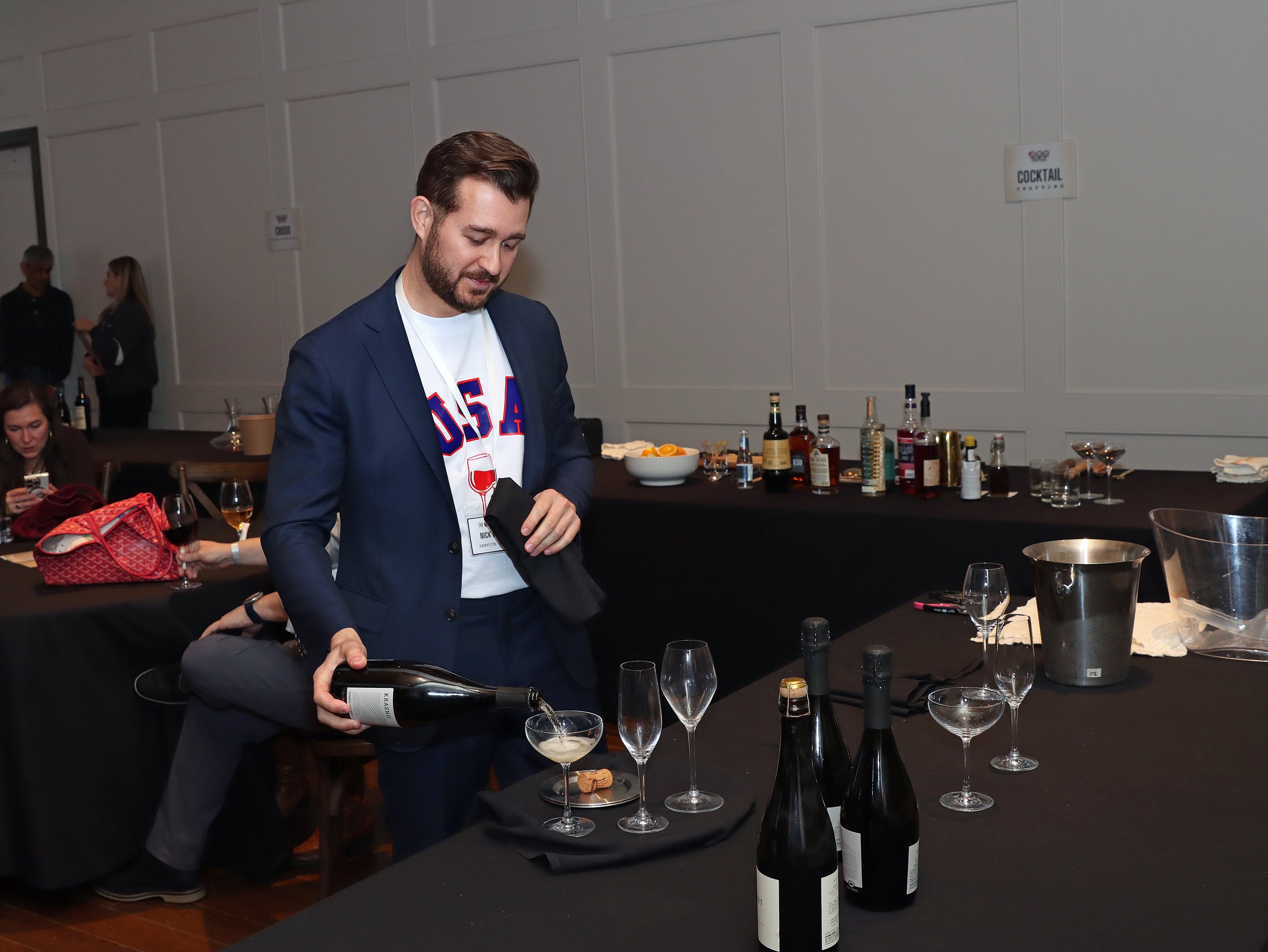 A man in a blue blazer and t-shirt with 'USA' on it is pouring red wine into a glass at a bar table with various bottles, glasses, and snacks in a social event setting.