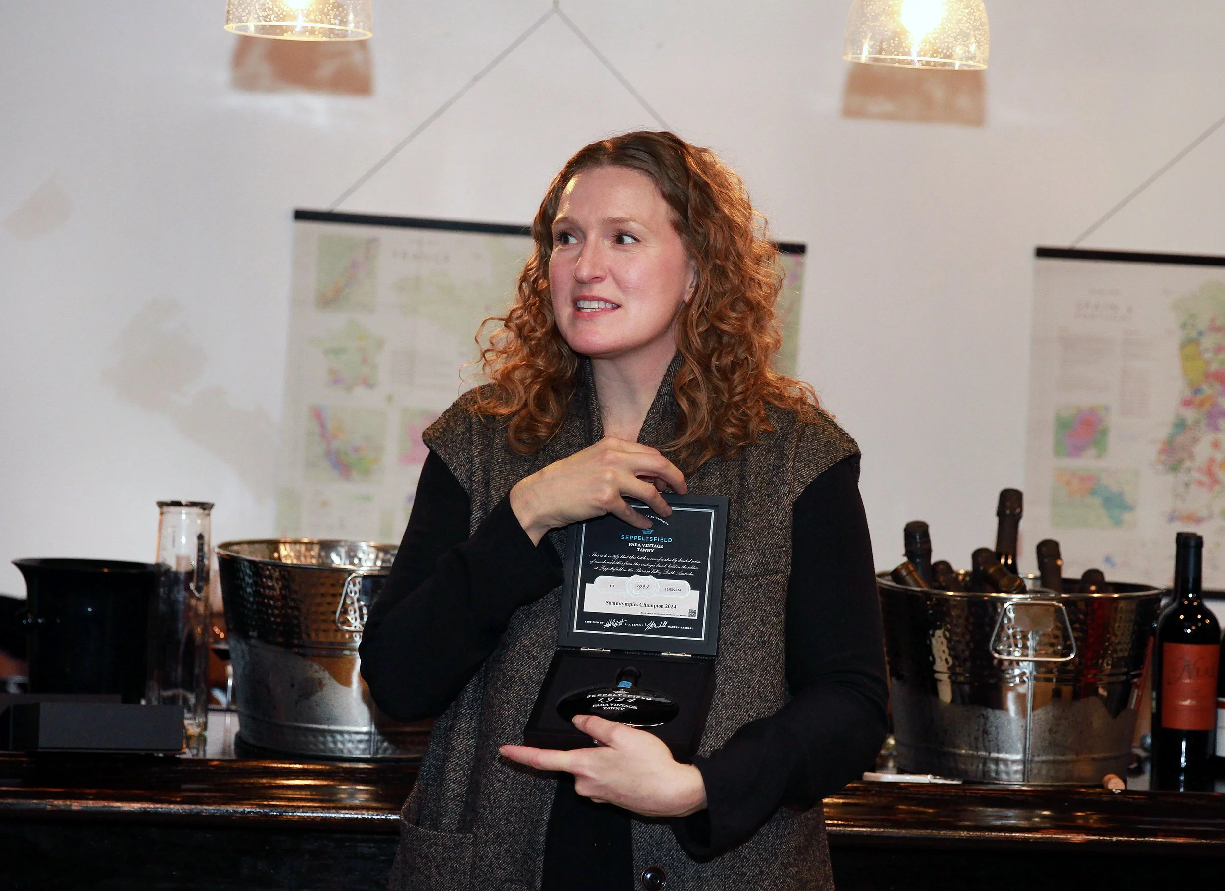 A woman with curly red hair holding an award, standing in front of a bar with wine bottles and glasses, with maps on the wall in the background.