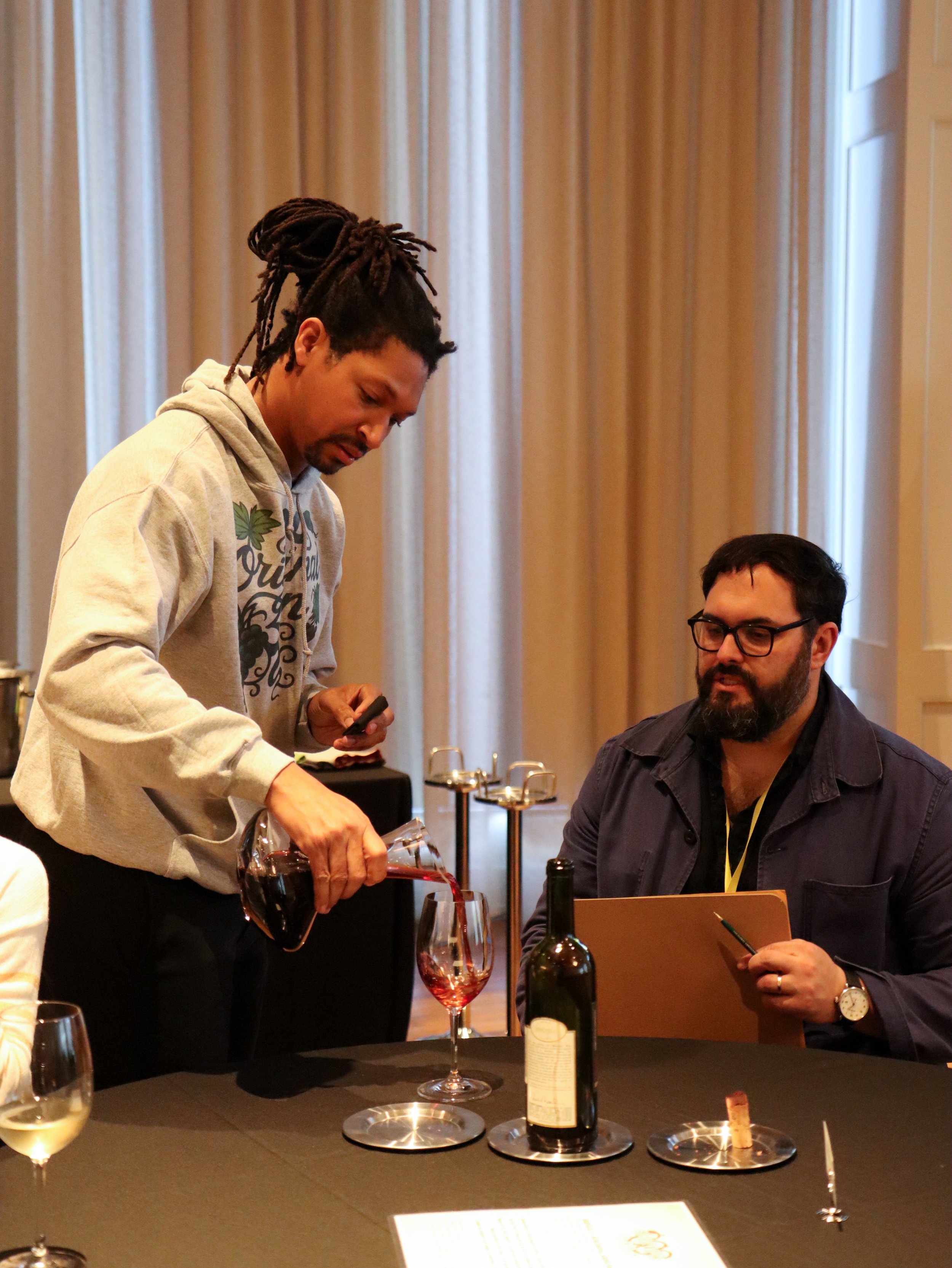 Two men engaging in tasting wine at a wine tasting event. One man is pouring wine into a glass while the other man is seated and holding a notebook. There are wine bottles, glasses with white wine and red wine, and a table setup visible.