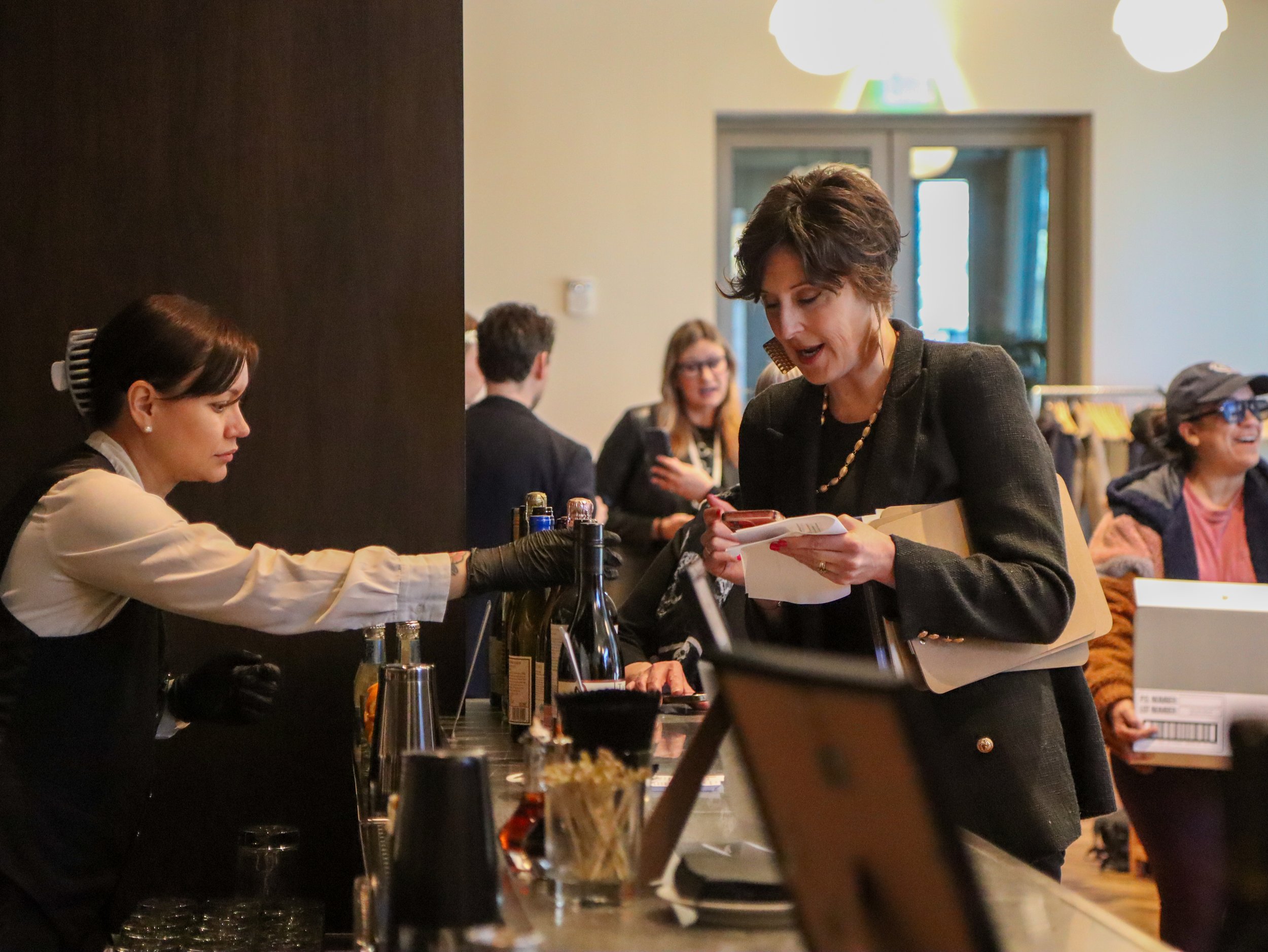 People at a bar or restaurant counter, engaging in conversation, with bottles, glasses, and bar tools visible on the counter, and patrons in the background.
