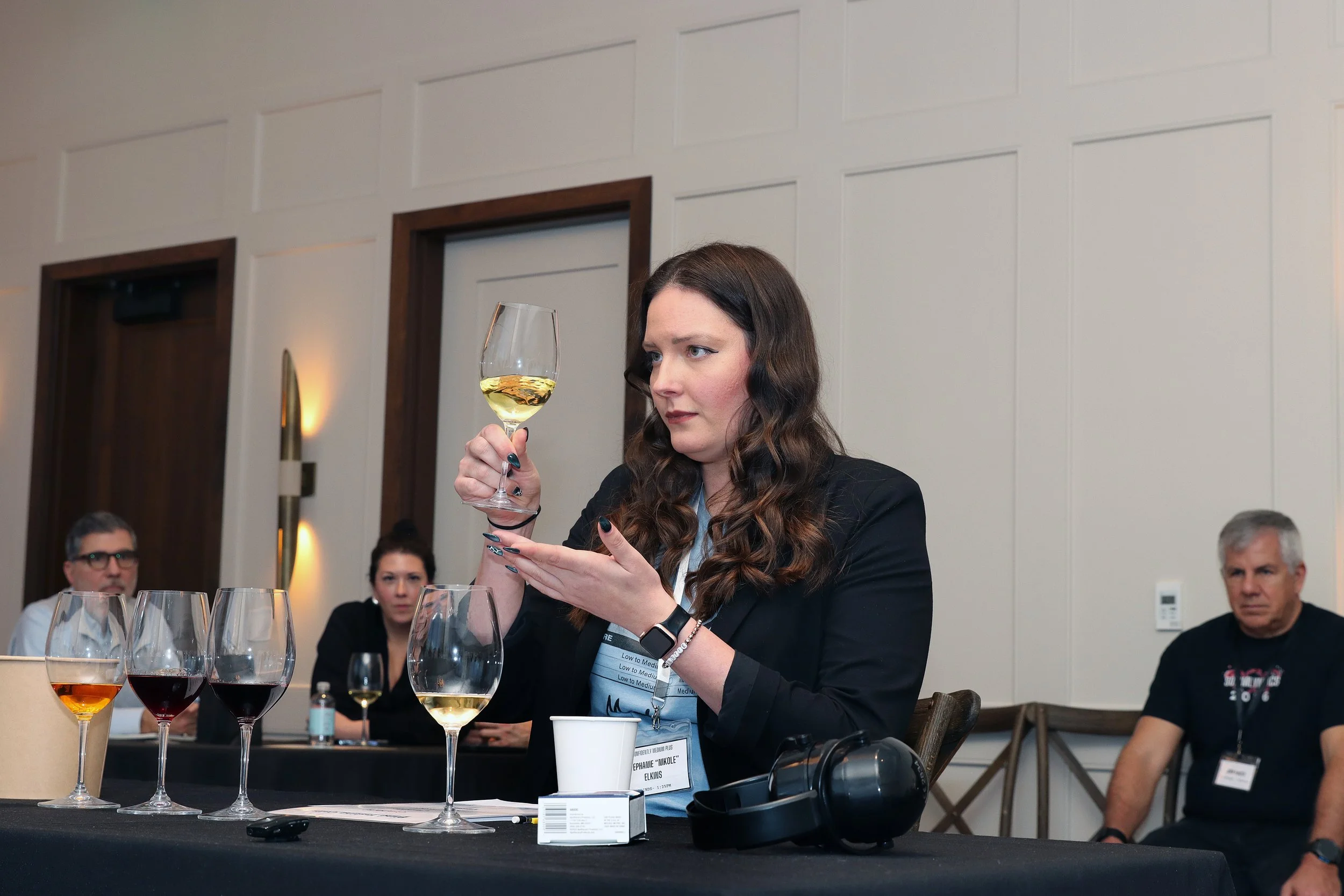 A woman with long dark wavy hair and a dark blazer is holding a glass of white wine, examining it closely during a wine tasting event. She is surrounded by other people, some seated and some standing, in a well-lit room with cream-colored walls and d