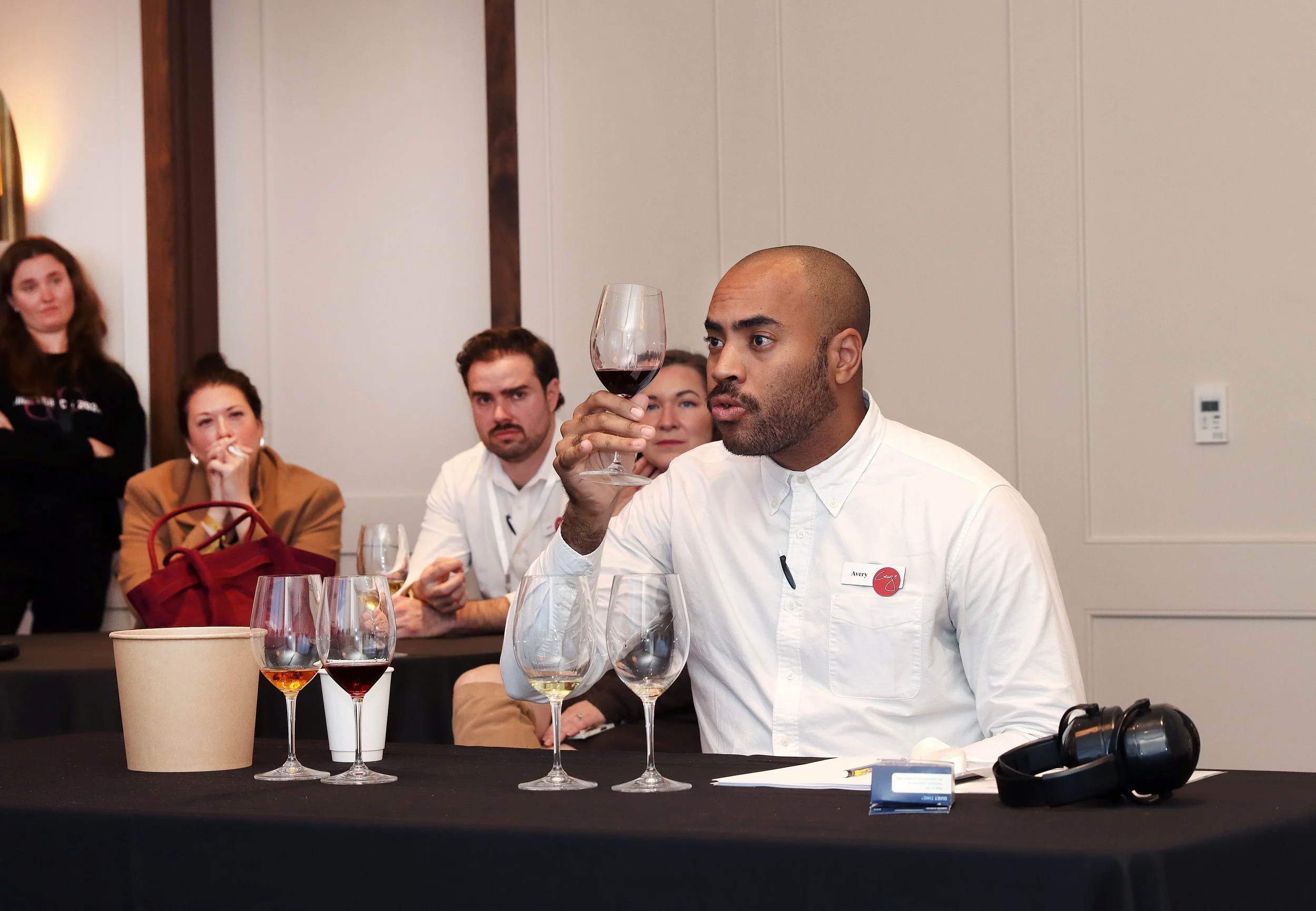 A man with a shaved head wearing a white shirt and a name tag tasting red wine at a tasting event, with three women sitting behind him observing, and multiple glasses and a paper cup on the table.