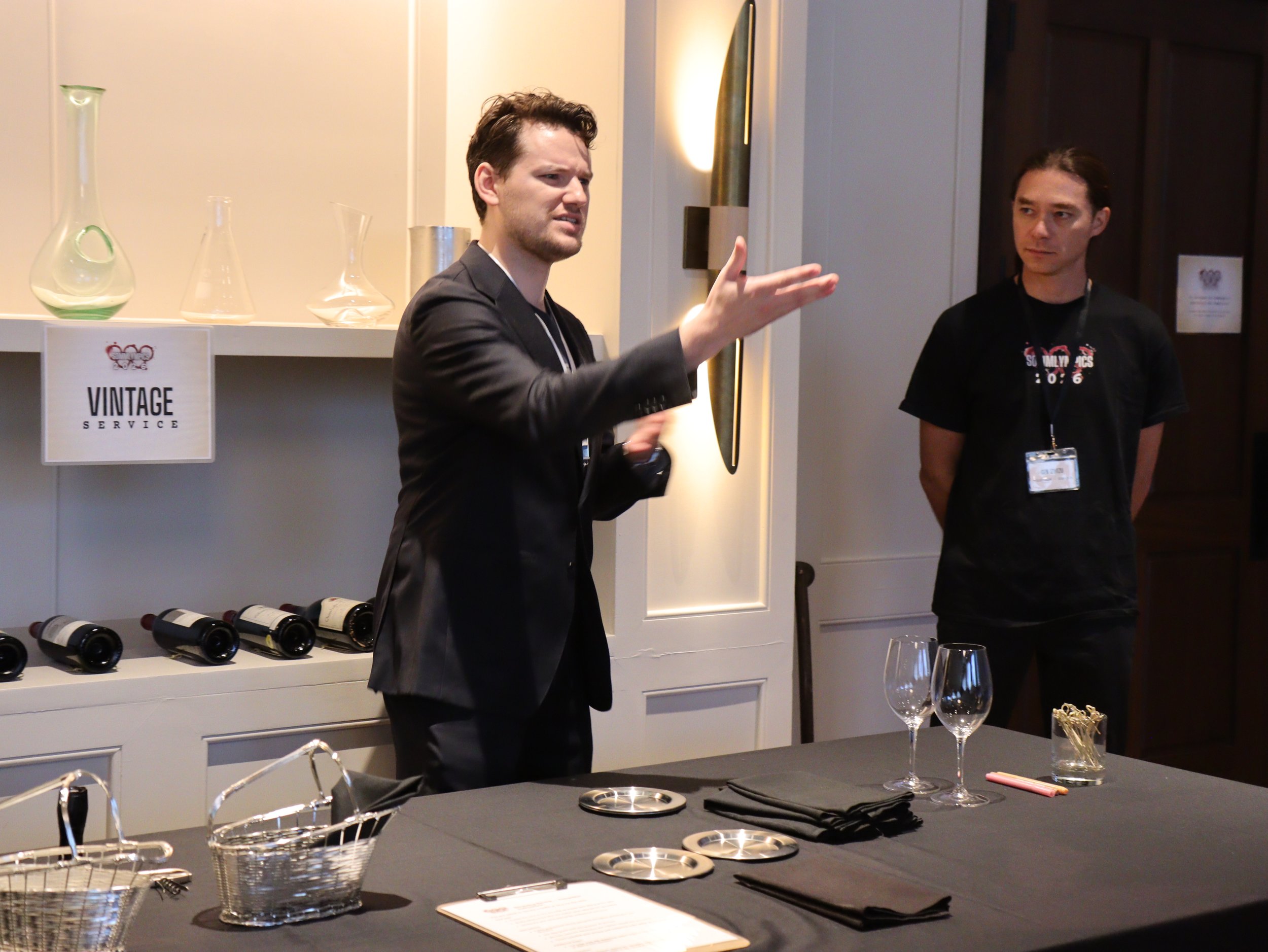 Two men are speaking at a wine tasting event, with wine bottles on a table and decorative glassware, in a room with a sign that says "Vintage Service" and wine glasses in the foreground.