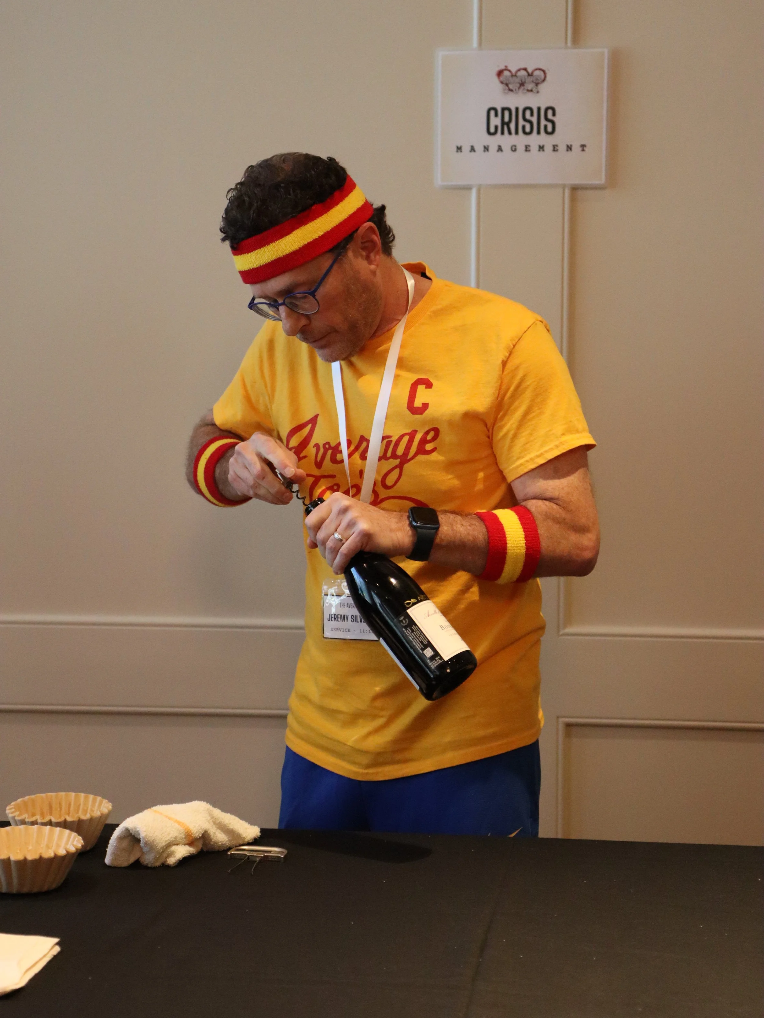 A man wearing a yellow T-shirt, red and yellow headband, and matching wristbands is opening a bottle of wine at a table with some cloth towels and paper cups. There is a sign on the wall behind him that reads 'Crisis Management'.