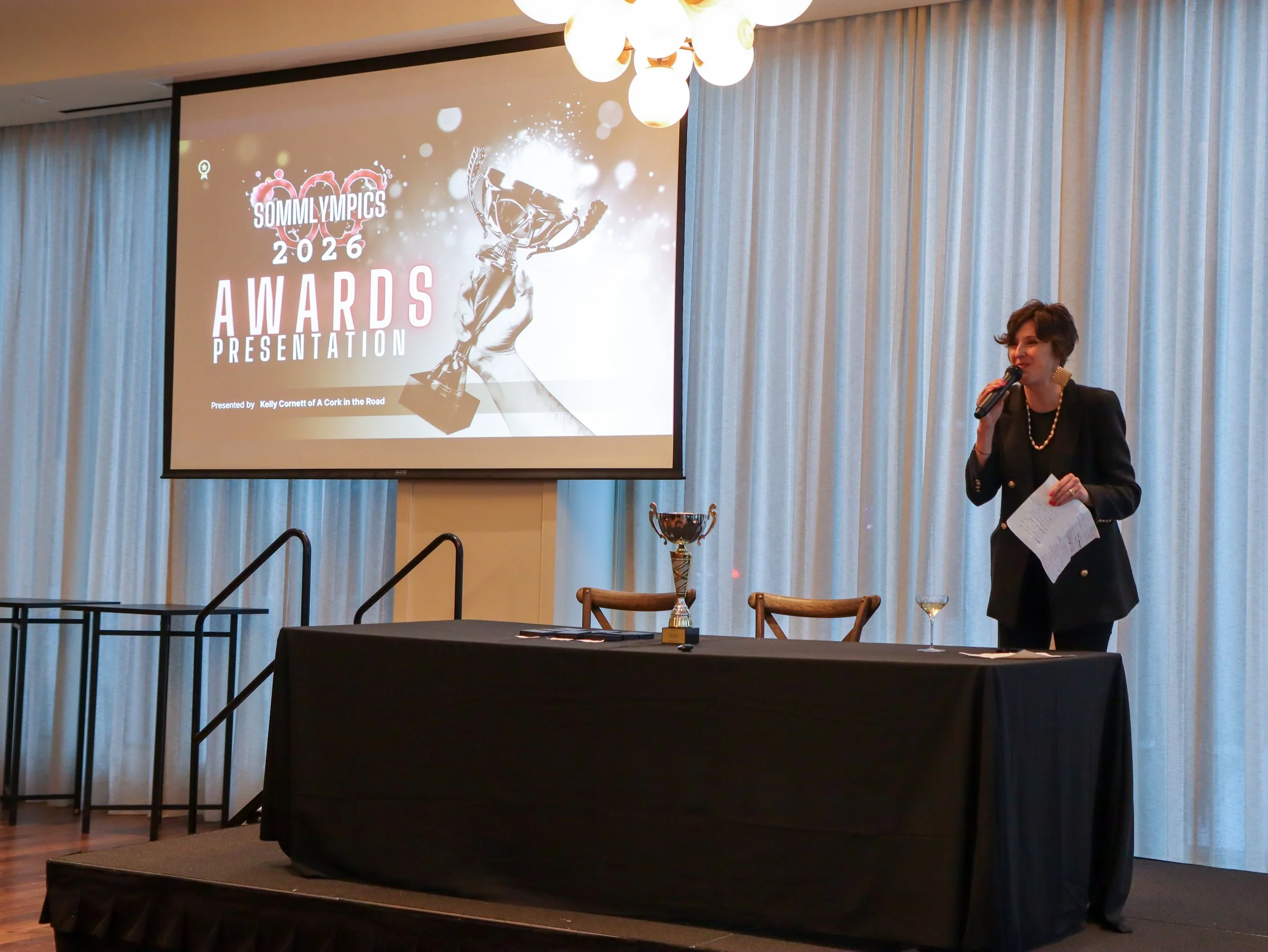 A woman is speaking into a microphone during an awards presentation at the SommyOlympics 2026 event, with a large screen behind her displaying the event logo and a trophy image.