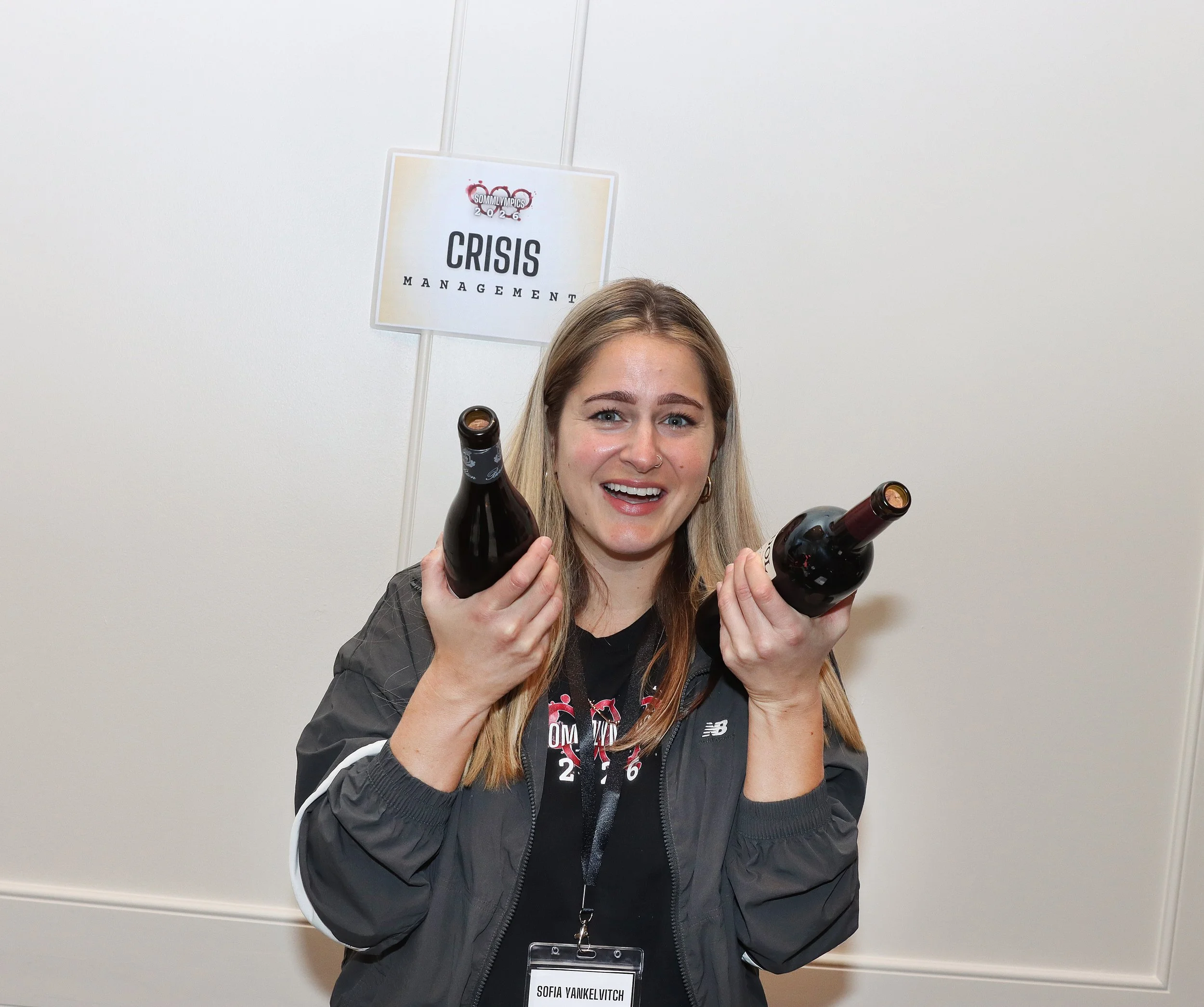 A young woman, smiling, holding two bottles of beer, standing in front of a plain white wall with a sign that reads 'Crisis Management'. She has a name tag that says 'Sofia Yankelvitch' and wears a black jacket and a lanyard.