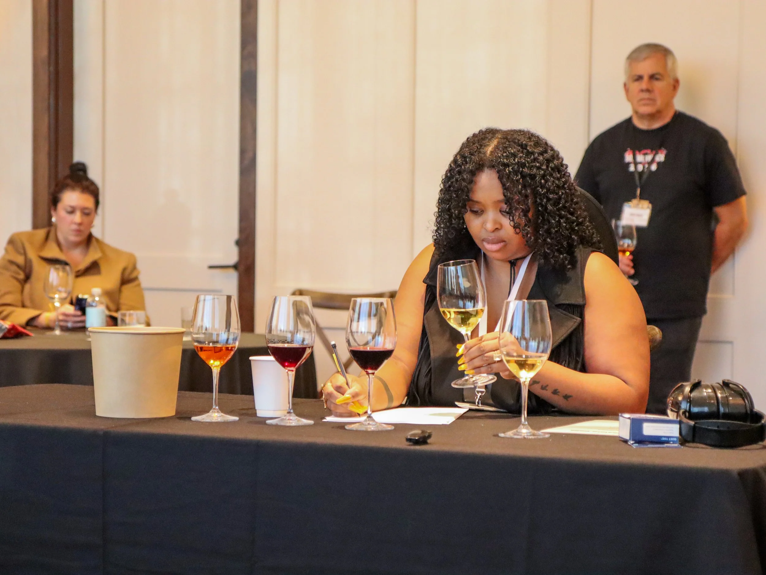 A woman with curly hair looking at a sheet of paper at a table with multiple glasses of white and red wine. A man in the background with glasses around his neck and a name badge, and another woman sitting on the left side.