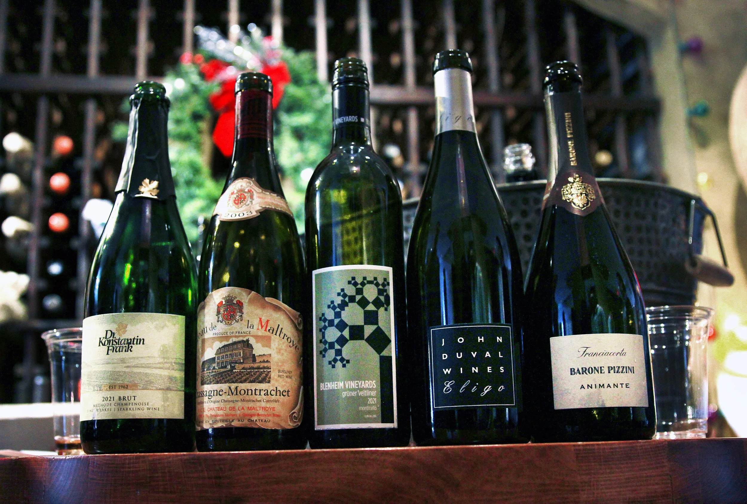 Five bottles of wine on a wooden surface with wine glasses, and a Christmas decoration in the background.