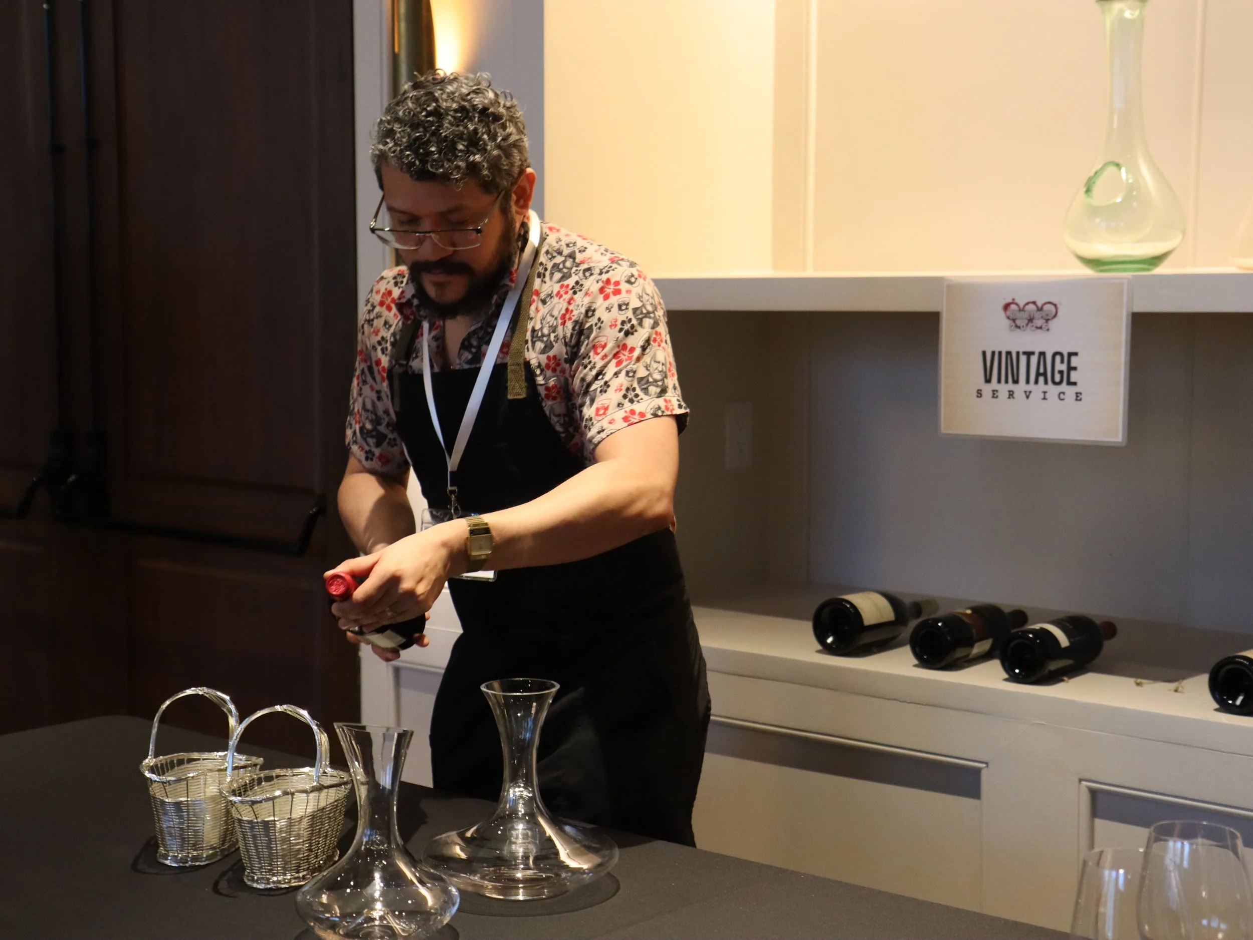Man with glasses and curly hair wearing a floral shirt and black apron preparing drinks at a bar or event, with wine bottles and a sign labeled "Vintage Service" in the background.