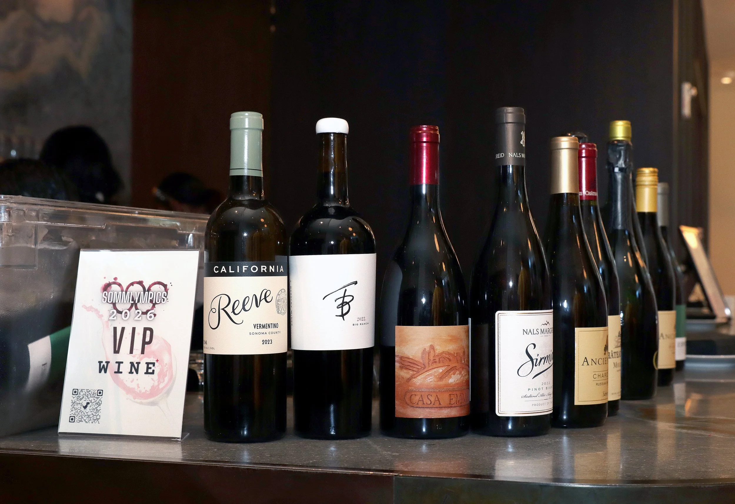 A row of wine bottles on a shelf with a sign that reads "SommyOlympics 2026 VIP Wine" and a QR code