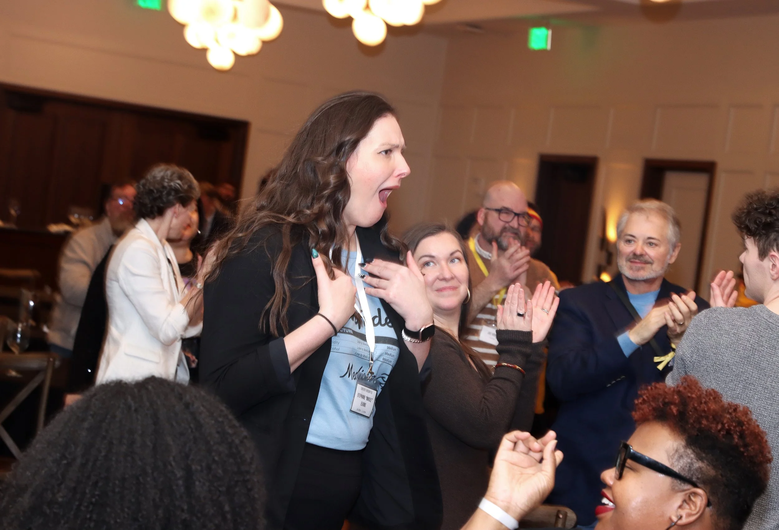 A woman with brown hair gesturing with her hands appears to be speaking or singing at an indoor event, surrounded by seated people clapping and smiling.