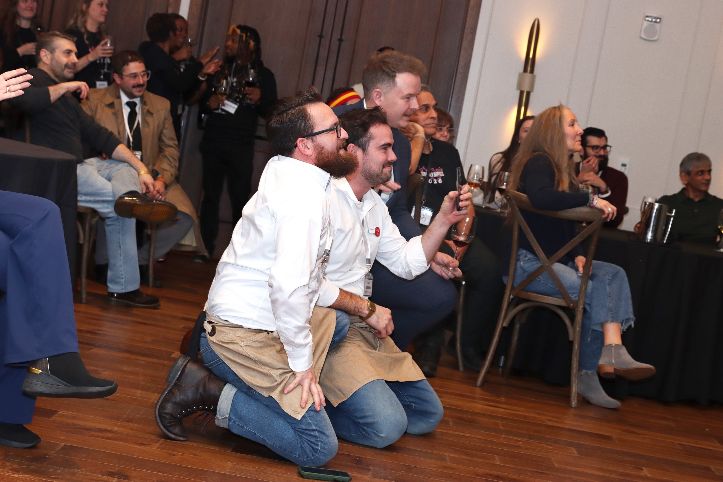 People at an indoor event, some are seated on the floor, others are sitting or standing at tables, enjoying and engaging with a presentation or performance, some holding drinks.