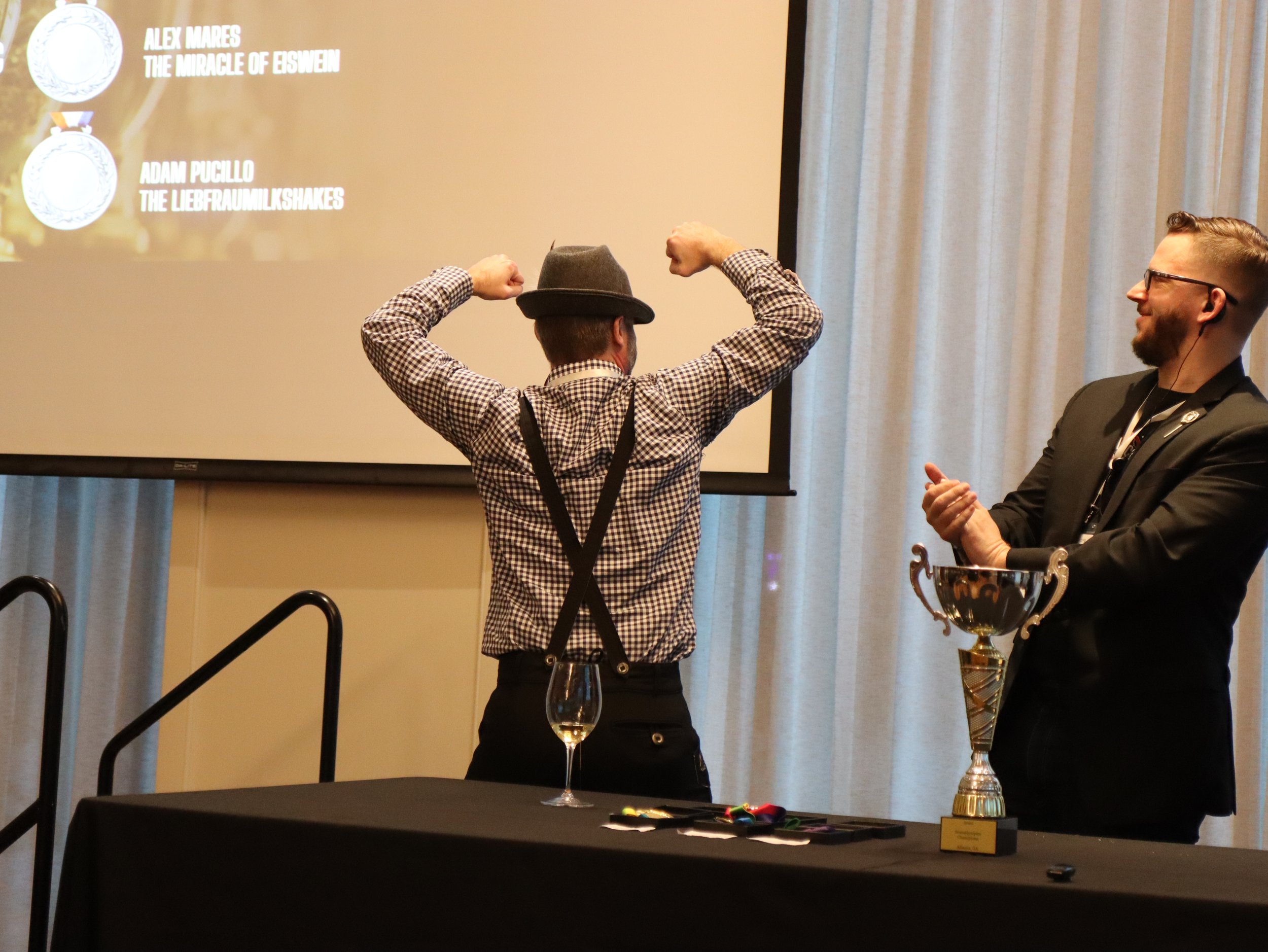 A man with a checkered shirt and hat flexing his arm muscles in front of a presentation screen. There is a man in a suit on the right with glasses and a beard, smiling. There is a table with a glass of wine, a trophy, and some ribbons or medals.