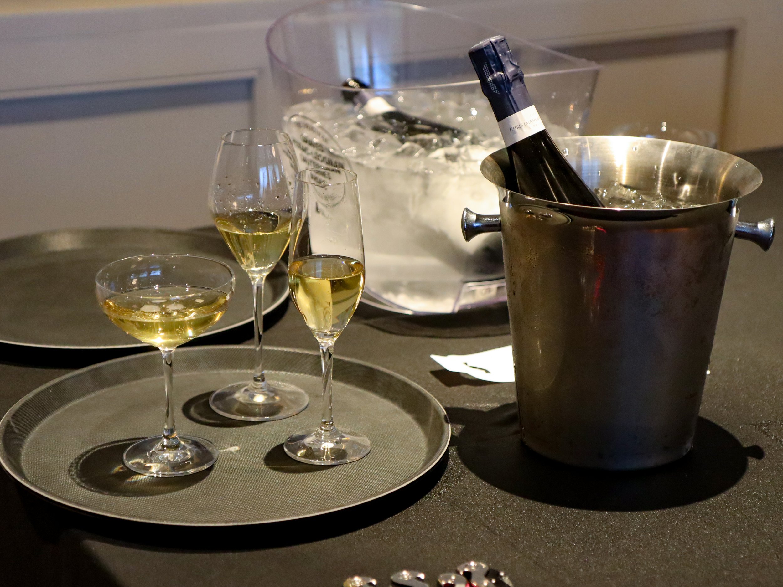 Three glasses of white wine on a silver tray, with ice and an ice bucket containing a bottle of wine in the background, placed on a dark countertop.