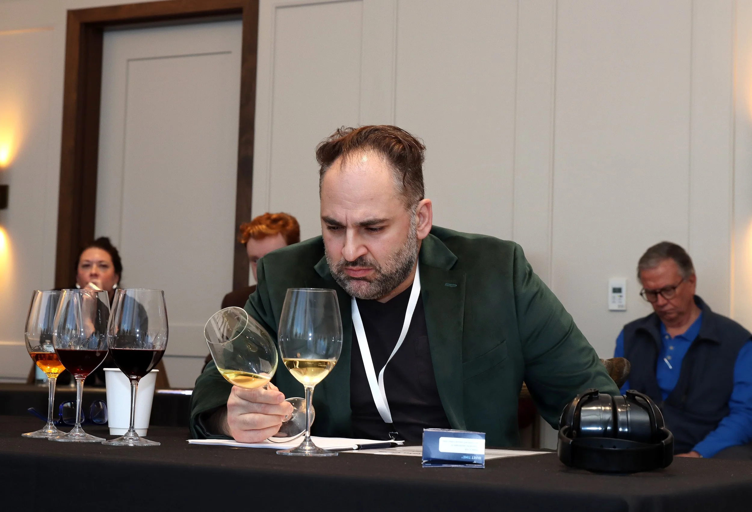 A man with a beard and dressed in a dark green blazer is tasting white wine in glasses at a tasting event, with other people in the background.