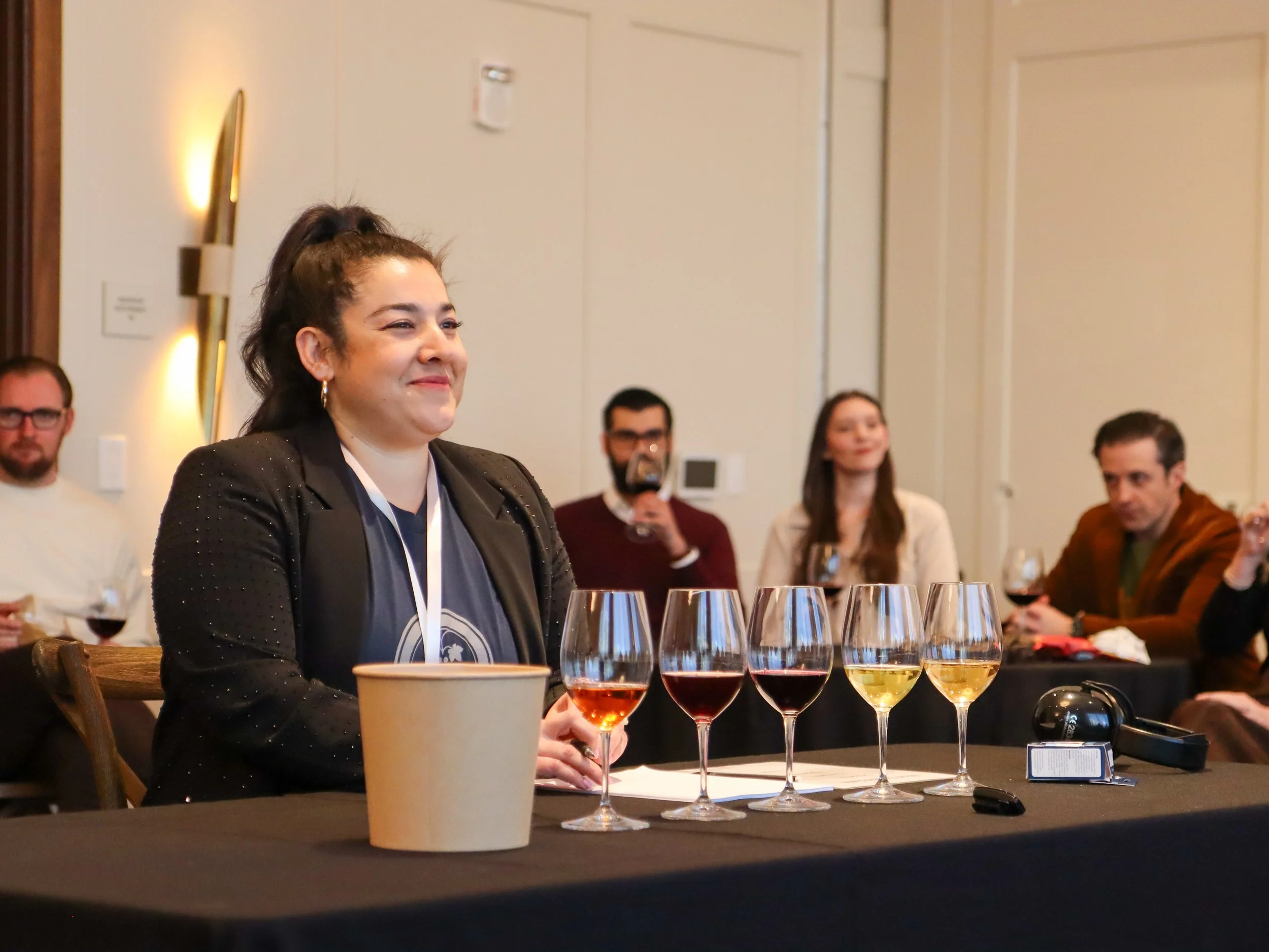 A woman with dark hair in a ponytail, wearing a black blazer with sparkles, seated at a table with a coffee cup and several glasses of wine or champagne in front of her, at an indoor event with other attendees in the background.