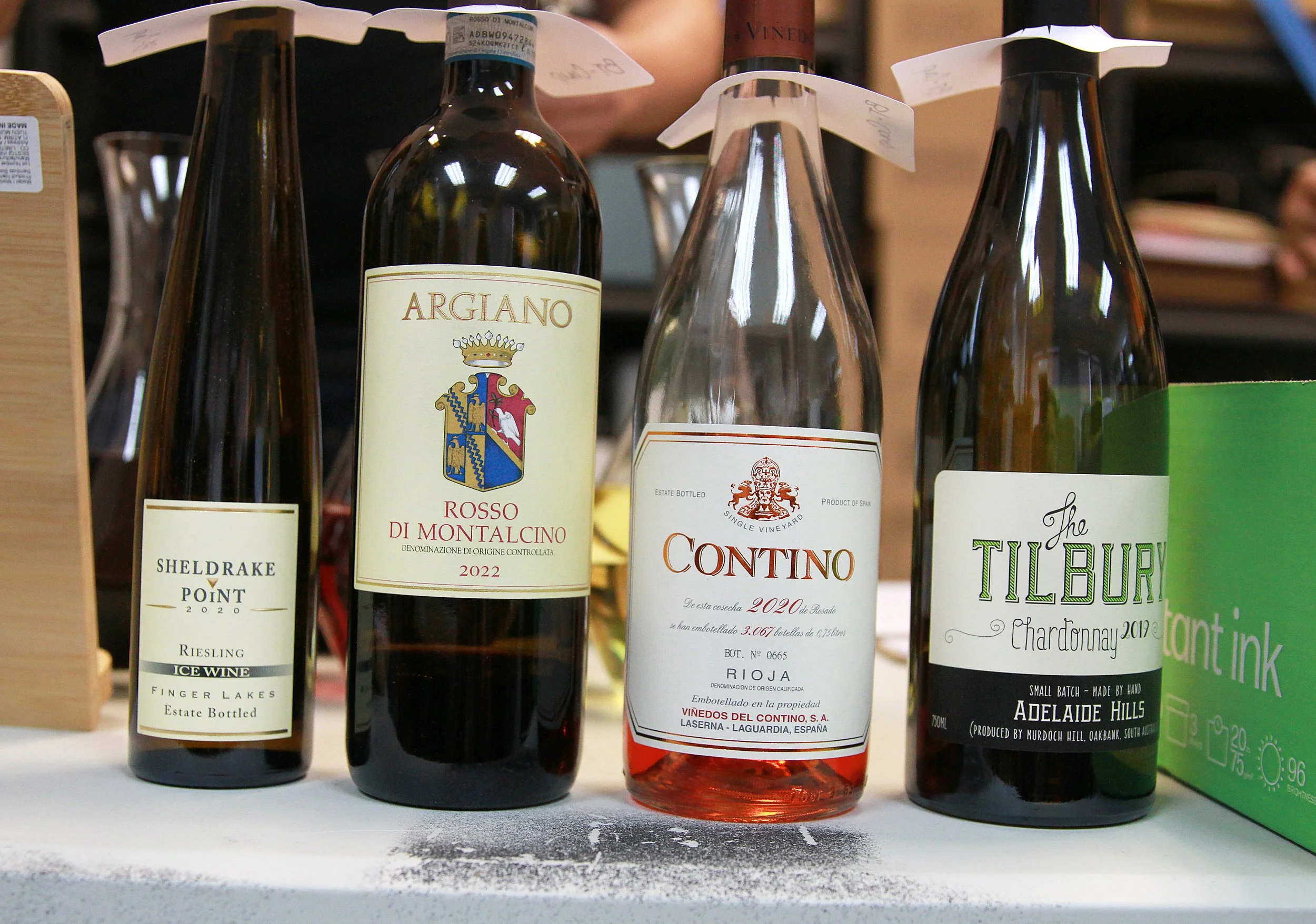 Four bottles of wine on a table, including a Riesling, a red wine called Argiano Rosso di Montalcino, a Rioja, and a Chardonnay. There are some green boxes on the right and a wooden sign on the far left.