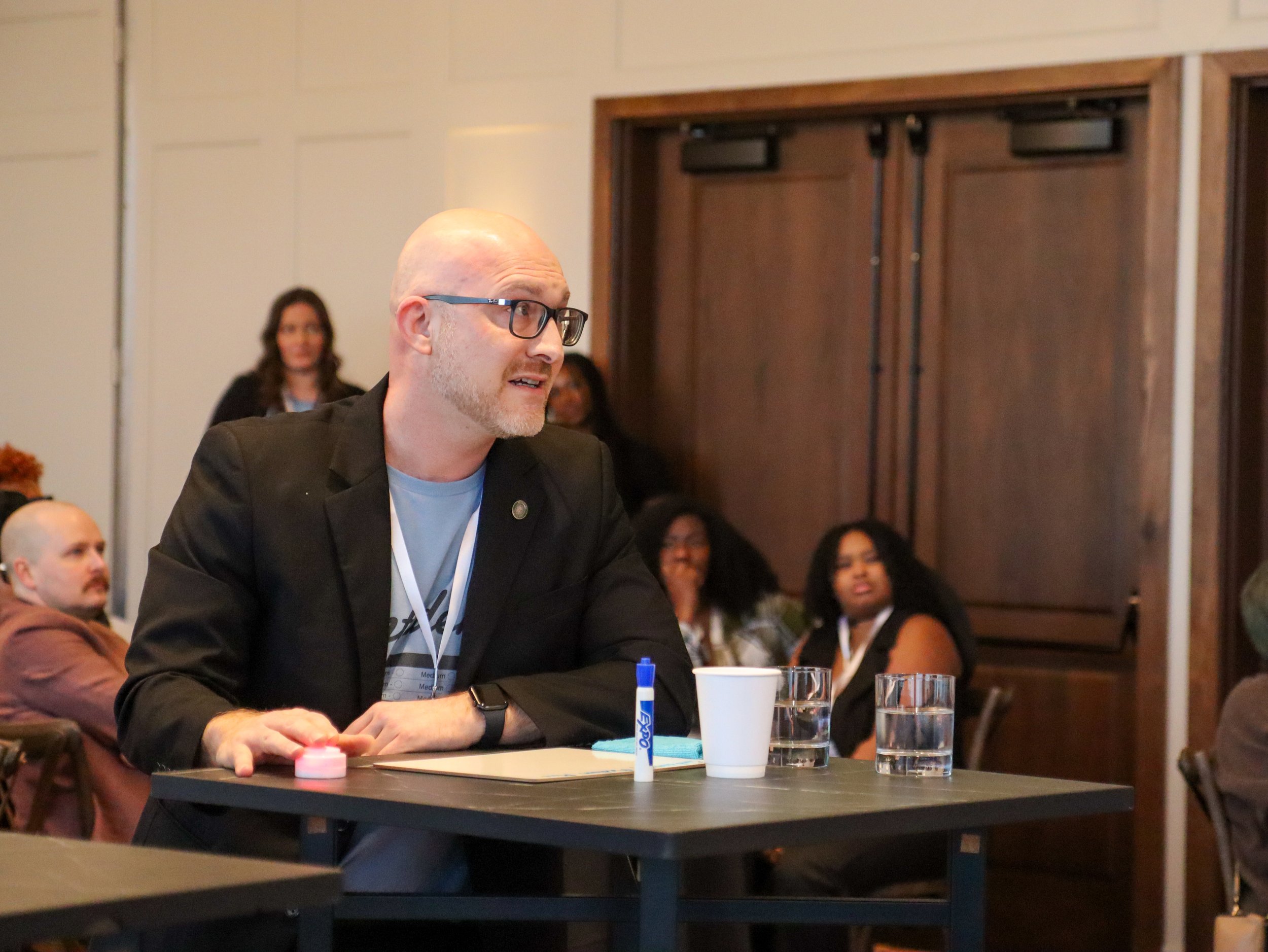 A man with glasses, a beard, and a bald head is sitting at a table during a meeting, with several glasses of water, a cup, a marker, and a small container in front of him. Other people are visible in the background, listening and smiling.
