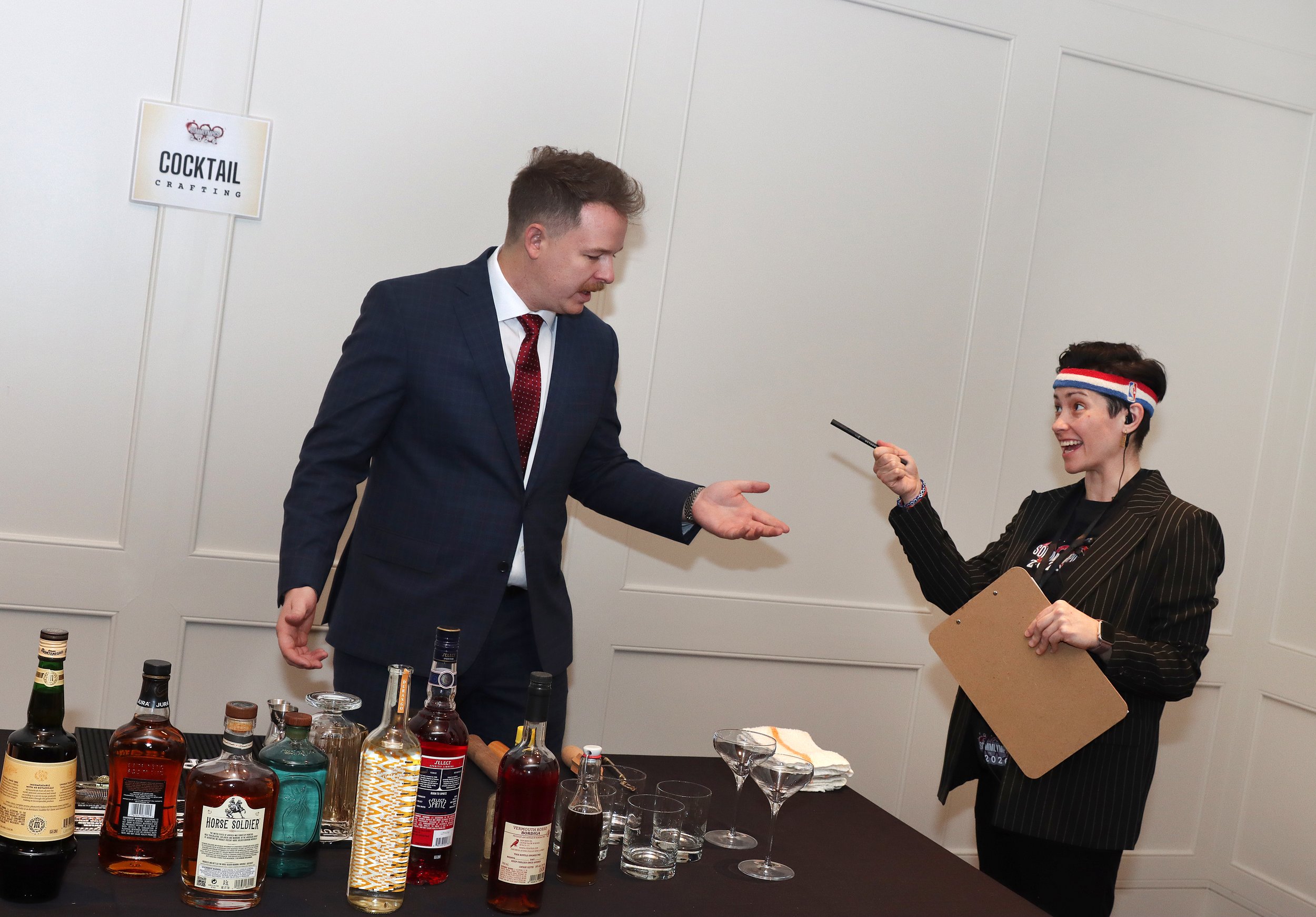 A man in a dark suit with a red tie is shaking hands with a woman wearing a striped blazer, a NBA headband, and holding a clipboard, in front of a table with various bottles of alcohol and empty glasses, at a cocktail crafting event in a room with wh