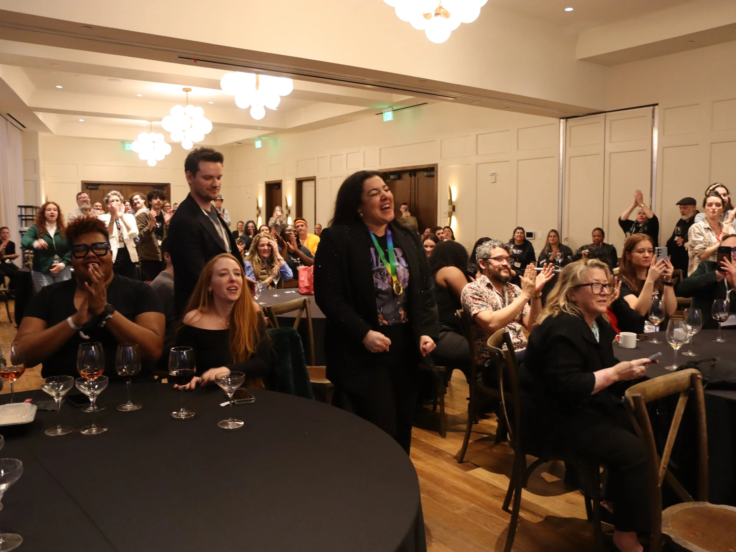 A group of people in a banquet hall attending a celebration, some are standing, some are sitting, smiling, clapping, and taking photos, with a woman in the center wearing a medal and a scrolling thankful expression.