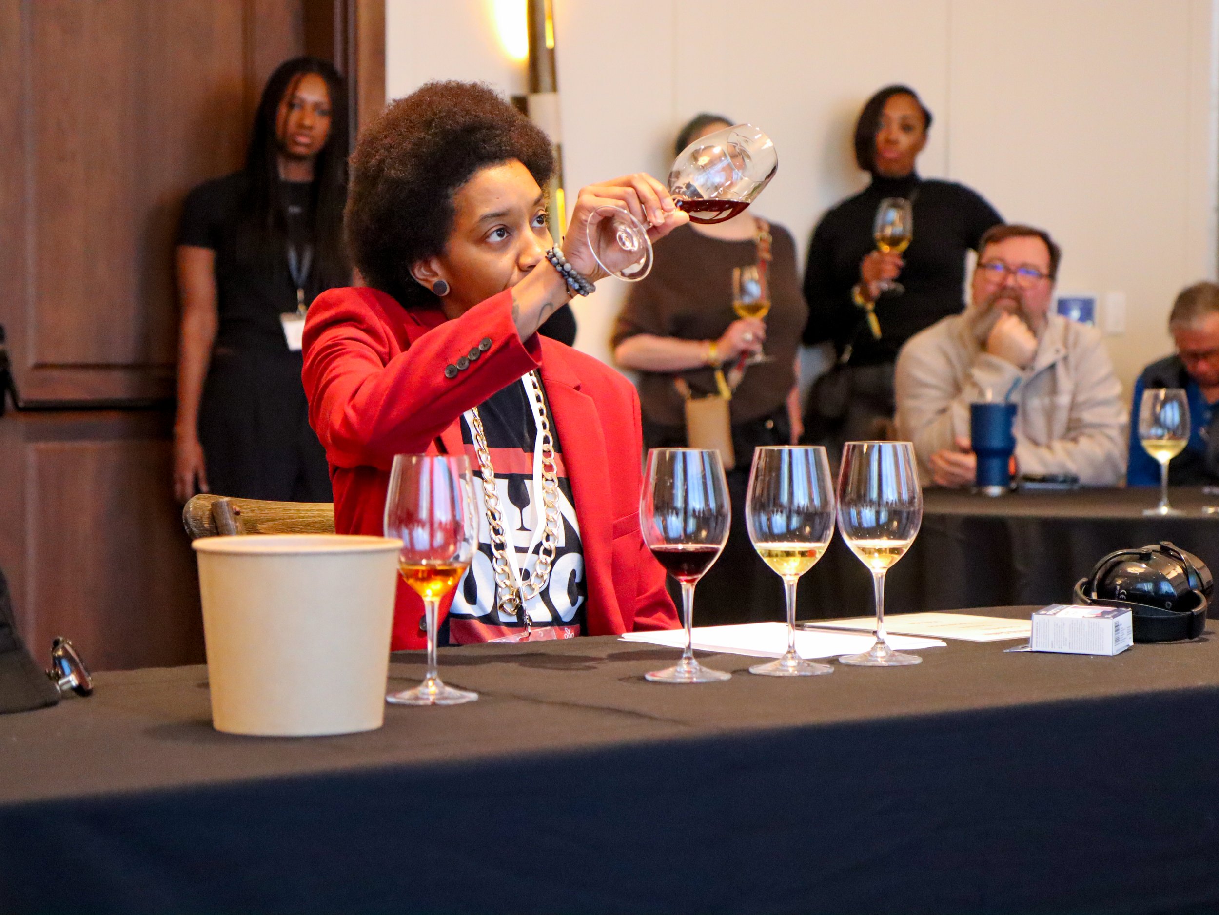 A woman in a red blazer and jewelry drinks wine at a tasting table with multiple glasses of wine in front of her. Several other people are in the background, some holding wine glasses, in an indoor setting.